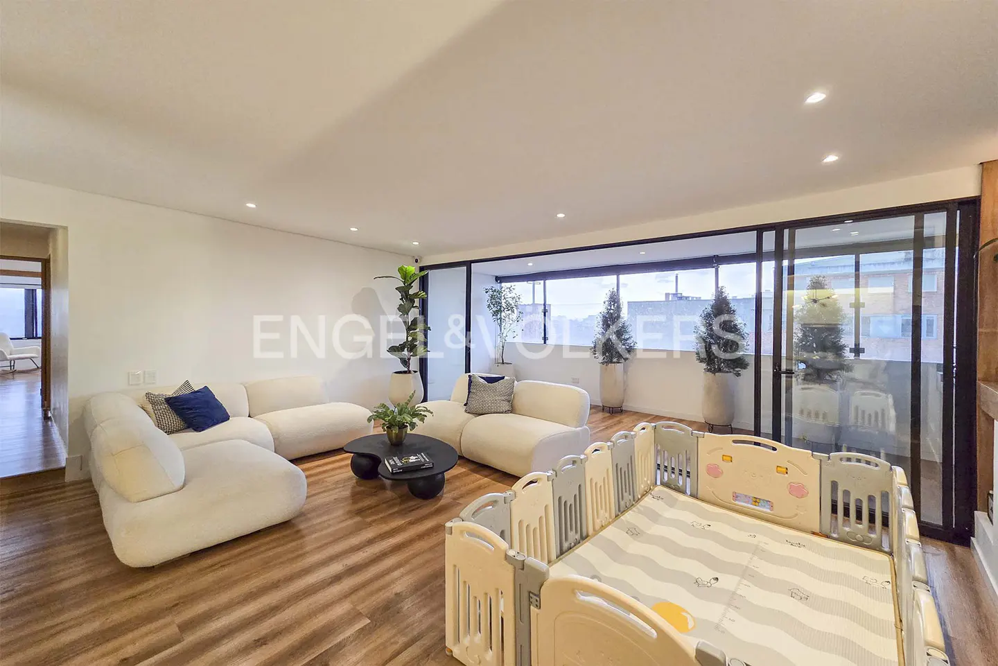 Bright living room with wood floors, white sofas, black table, and a baby playpen. Large windows offer city views.