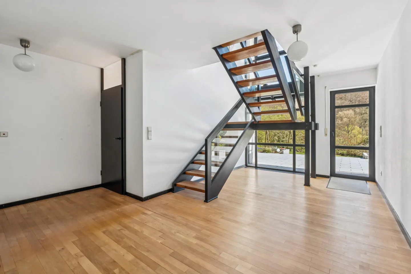 Bright, modern foyer with hardwood floors, white walls, and a black metal staircase with wood treads and glass railings. A black door and glass doors lead outside.