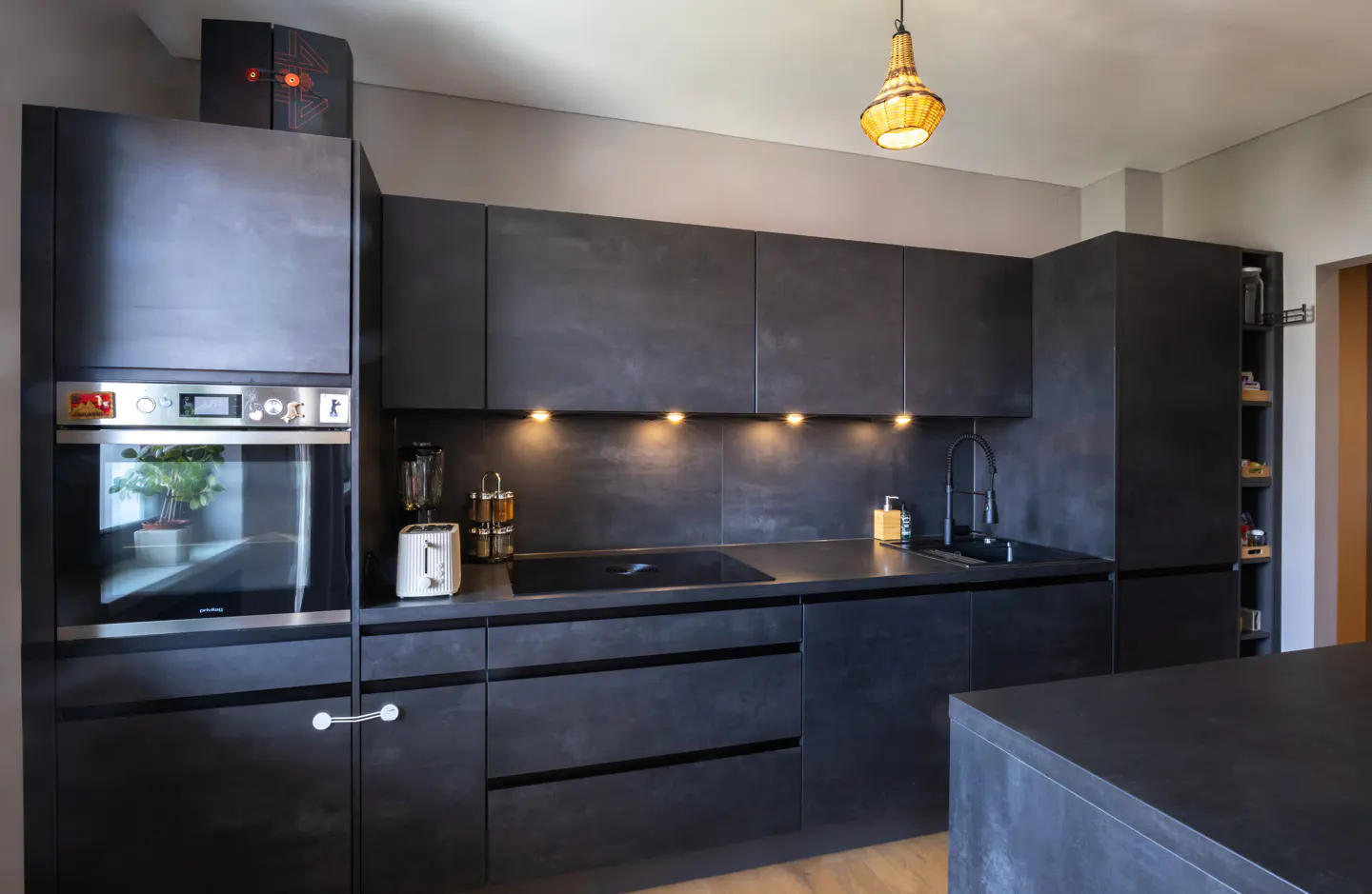 Modern kitchen with dark gray cabinets, black countertops, and stainless steel appliances. A woven pendant light hangs above.