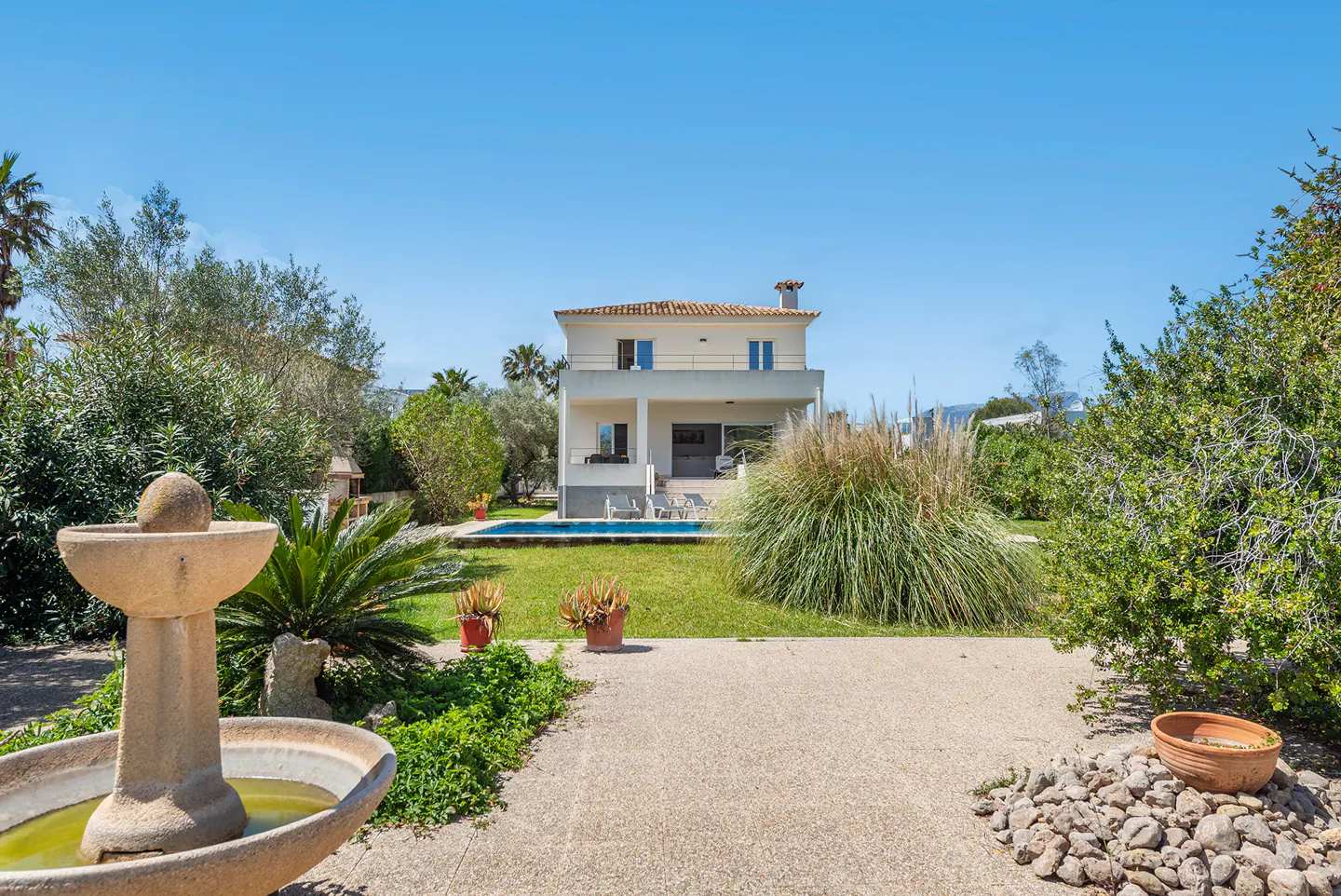 Two-story house with a pool and lush landscaping under a blue sky. A stone fountain is in the foreground.