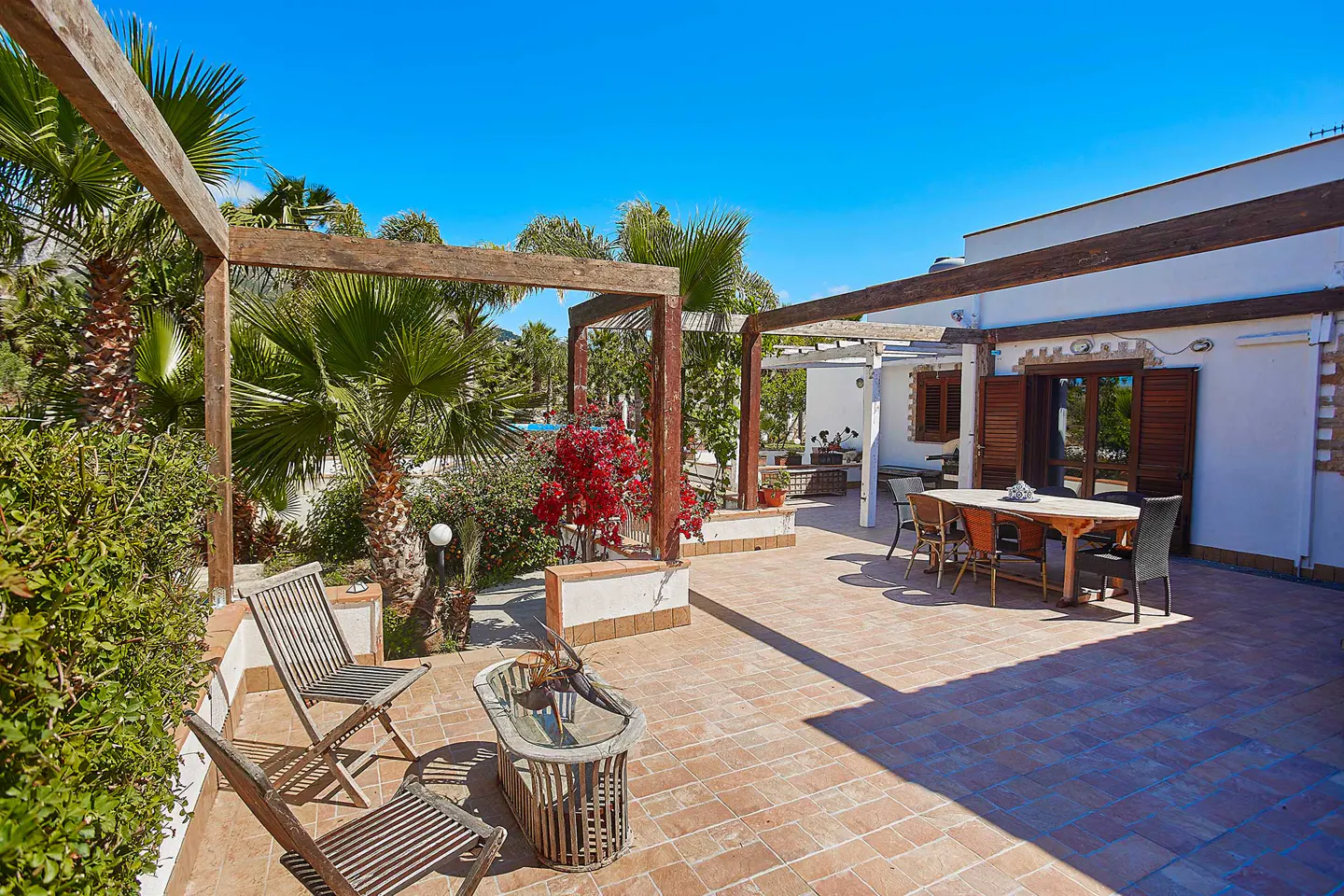 Sunny outdoor patio with terracotta tiles, wooden pergola, dining set, and lush palm trees against a clear blue sky.
