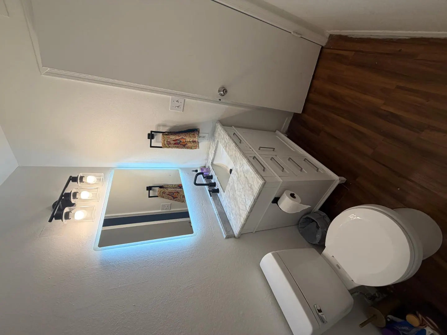 Bathroom with white walls, wood floor, toilet, vanity with marble top, and a mirror with blue LED lighting.