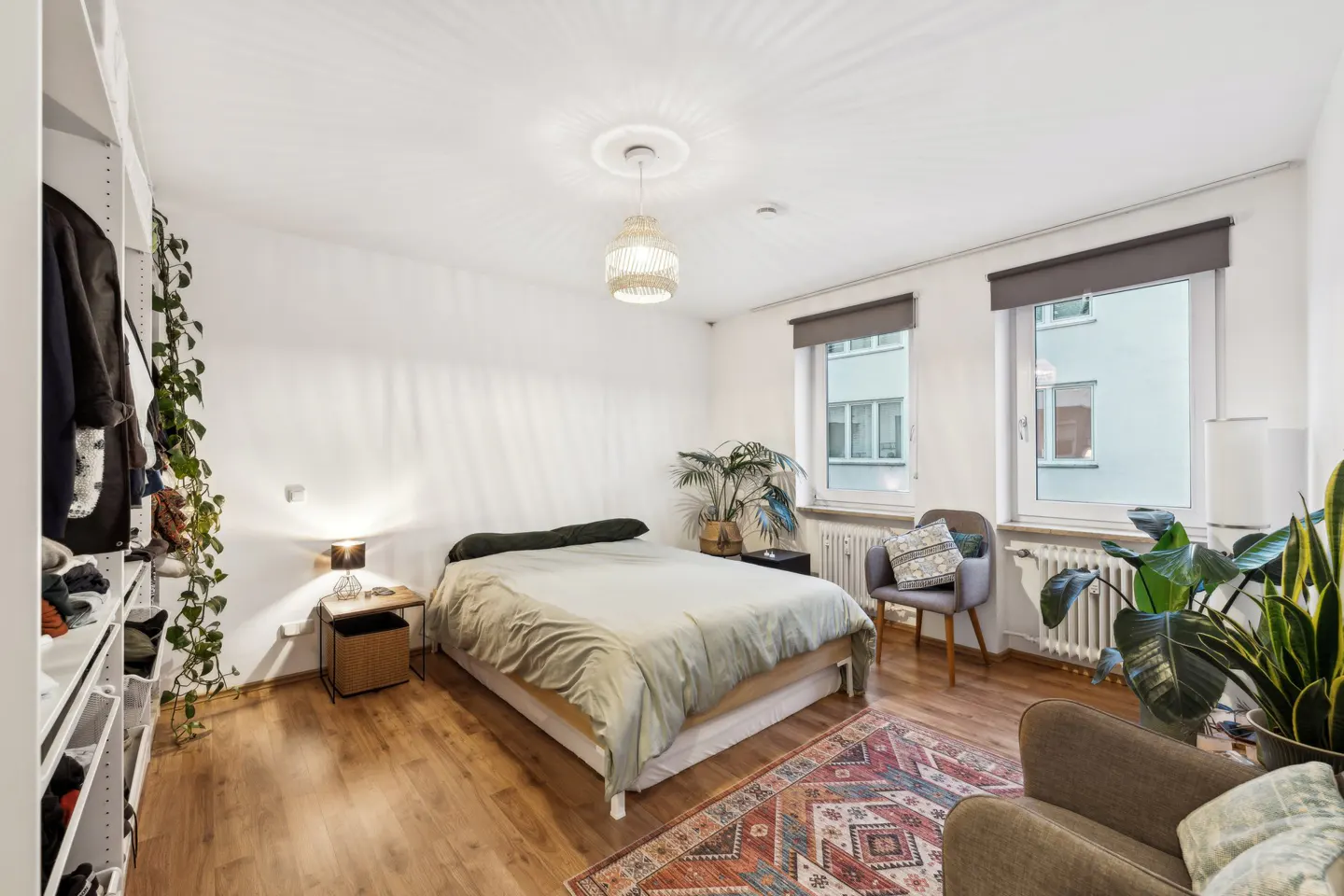 Bright bedroom with wood floors, white walls, and two windows. A bed, chair, and armchair are present, along with plants and a patterned rug.