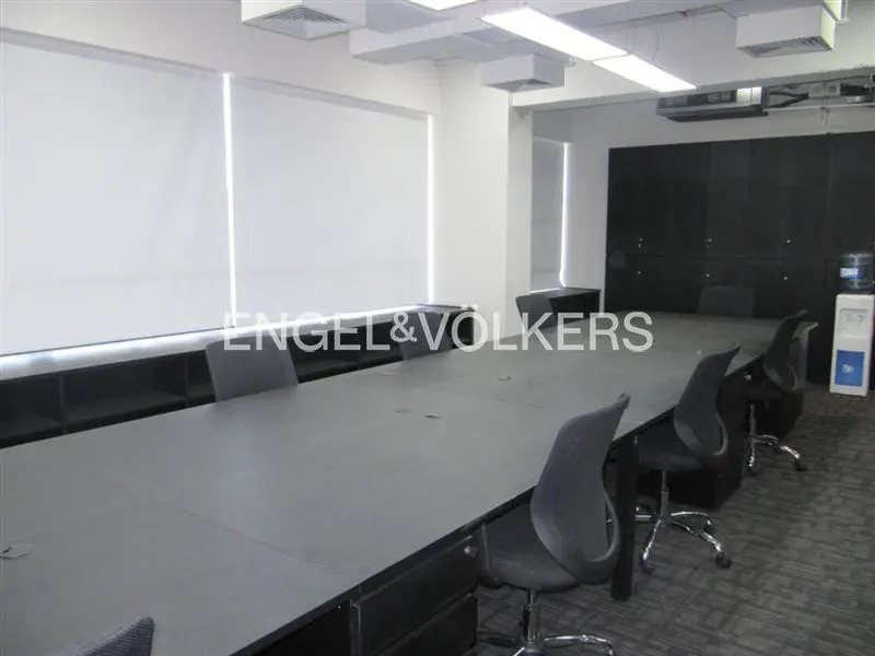 A modern office space with a long conference table and black chairs. White blinds cover the windows, and a water cooler stands in the corner.