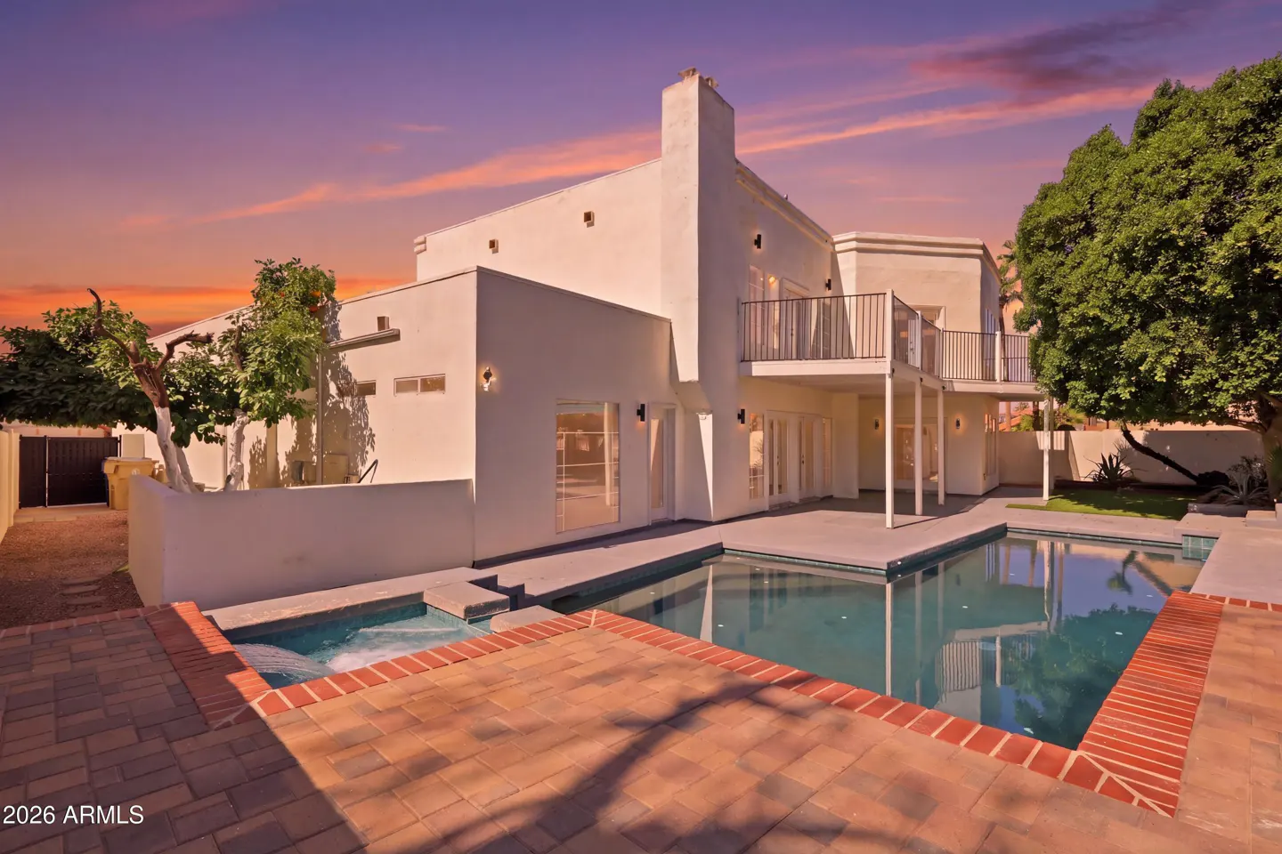 Exterior view of a two-story white house with a pool and spa, brick patio, balcony, and trees against a colorful sunset sky.