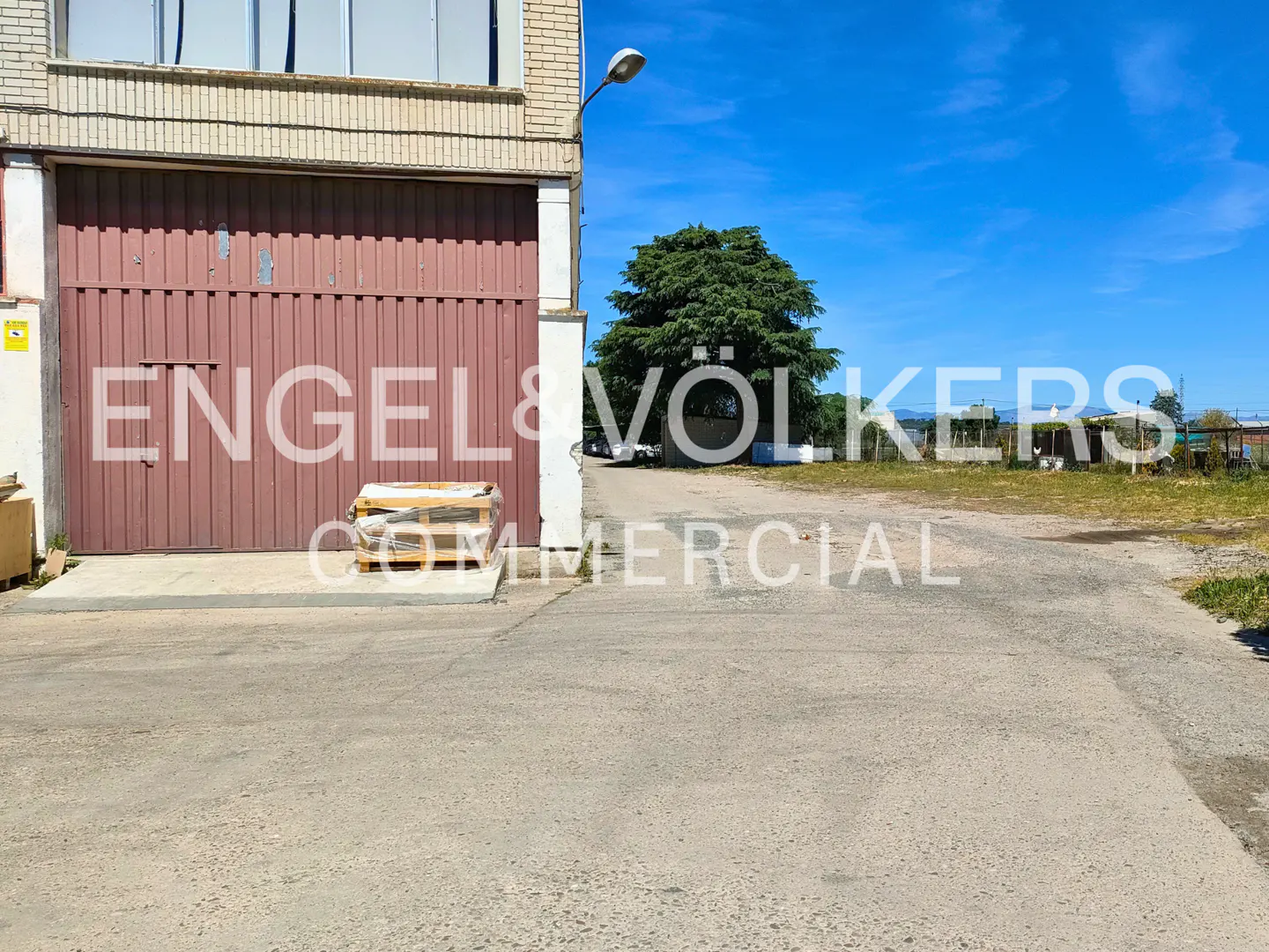 Exterior view of a commercial building with a large brown metal door, a wooden pallet, and a paved lot under a blue sky.