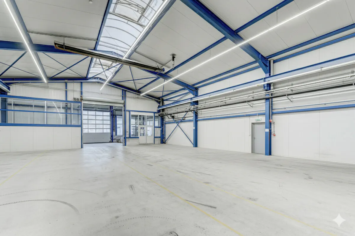 Bright, empty warehouse with white walls, gray floor, and blue steel beams. Skylights and LED lights illuminate the space. A large garage door is partially open.