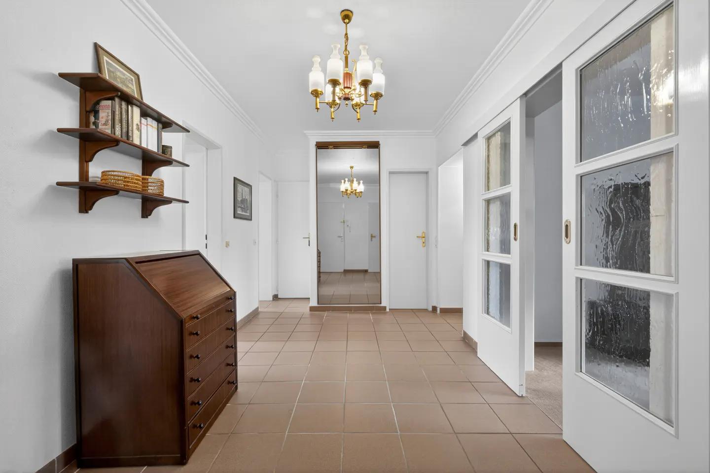 Hallway with brown tile floor, white walls, and a vintage desk. A gold chandelier hangs from the ceiling.