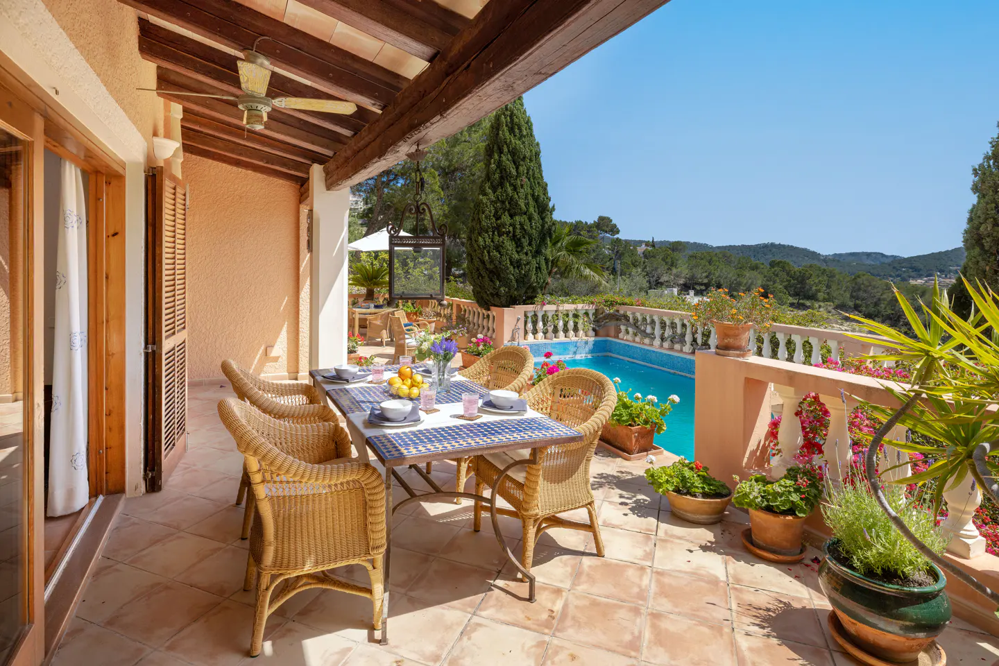 Outdoor patio with a table set for a meal, wicker chairs, and a pool with a view of trees and a blue sky.