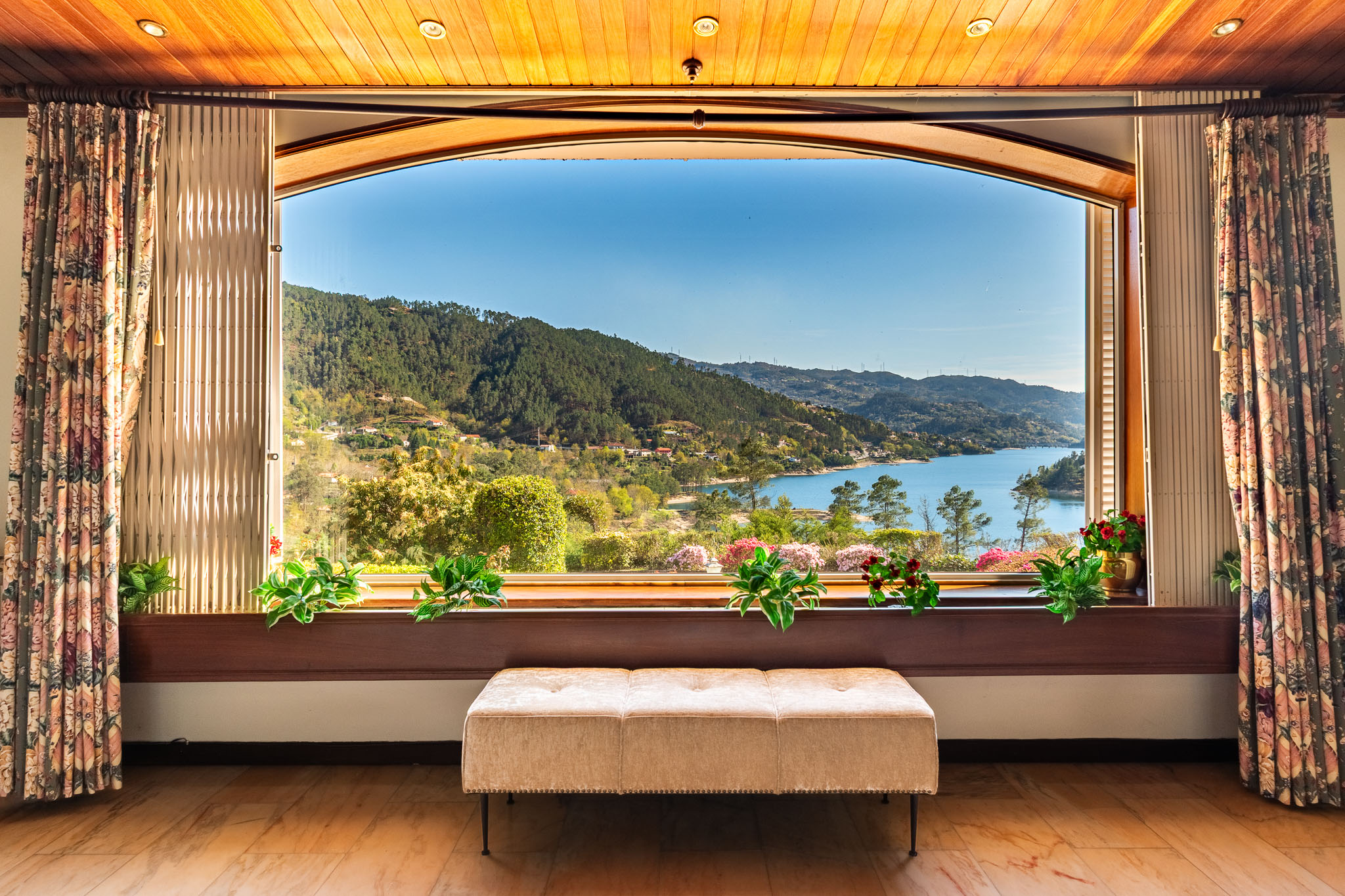 Interior view of a room with a large arched window showing a lake and mountains. Floral curtains frame the window, and a beige bench sits in front.