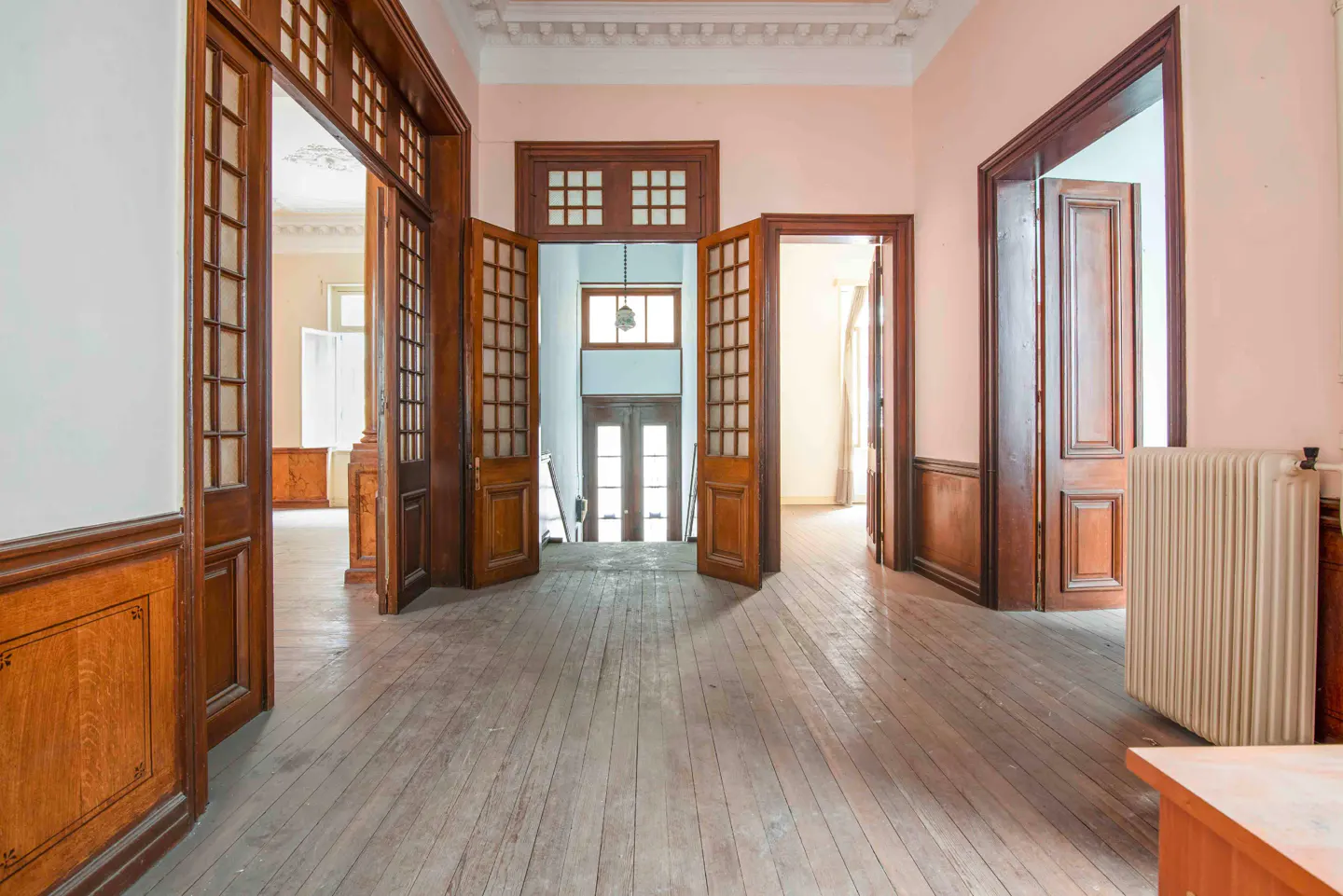 Hallway with wood floors, pink walls, and multiple open wood doors leading to other rooms. A radiator is visible on the right.