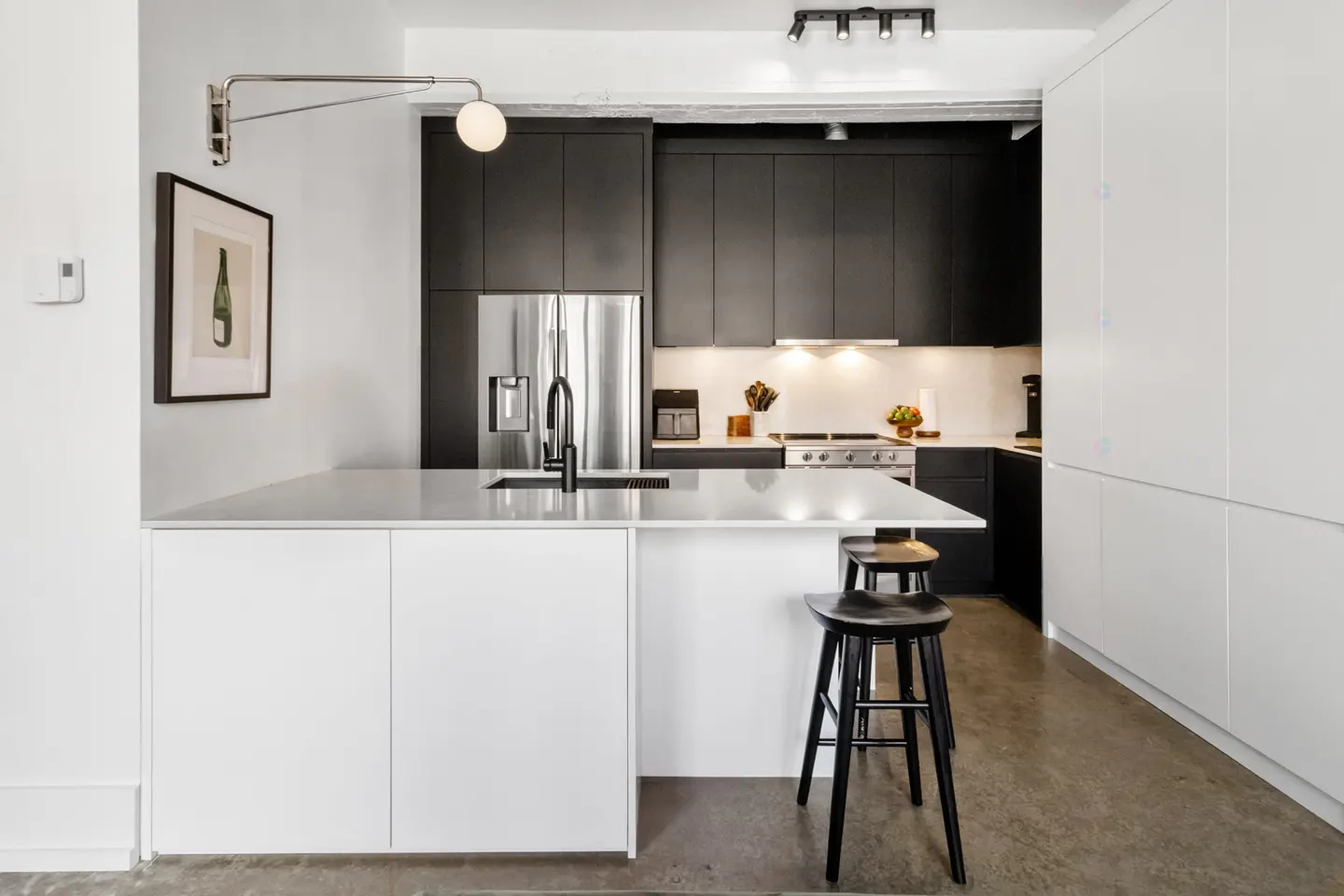 Modern kitchen with black cabinets, stainless steel fridge, white island with stools, and a framed art print.
