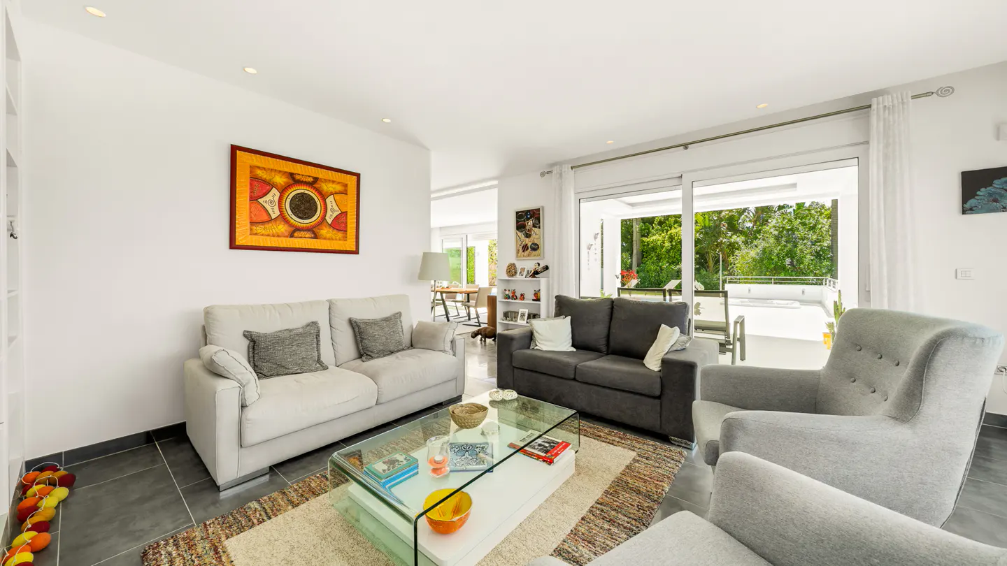 Bright living room with a gray sofa, a light gray sofa, and two light gray armchairs. A glass coffee table sits on a patterned rug. Sliding glass doors lead to a patio.