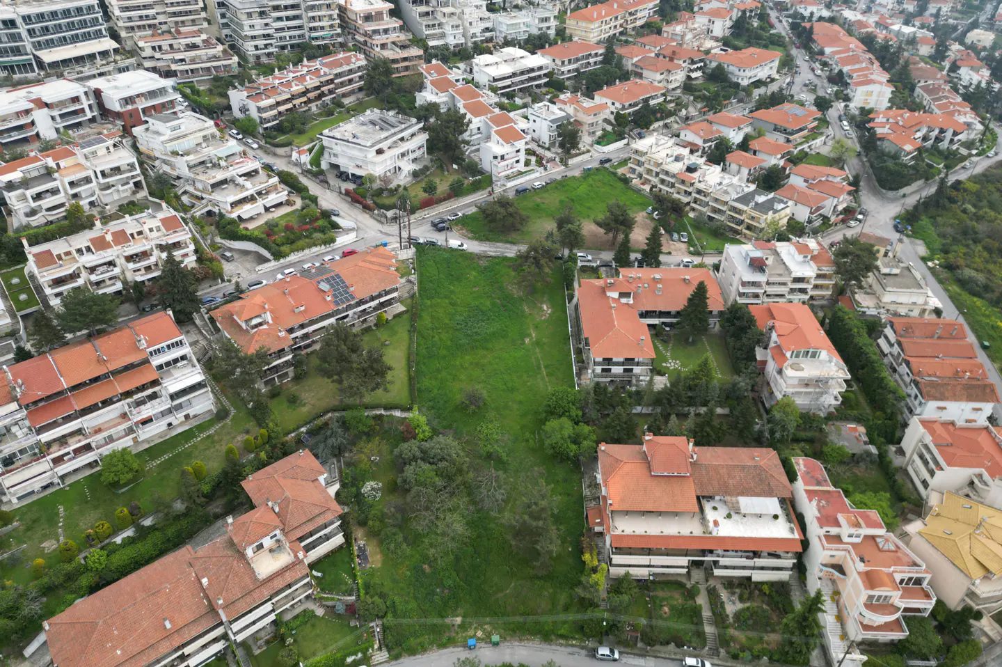 Aerial view of a green lot surrounded by buildings with orange tile roofs in a residential area.