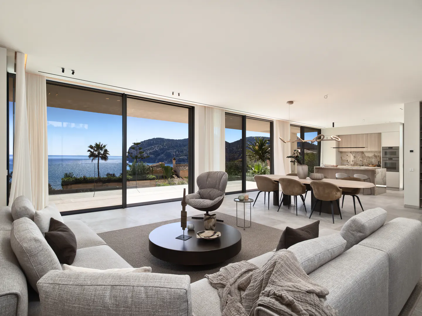 Bright, modern living room with a gray sectional sofa, round coffee table, and ocean view through large windows. Dining area and kitchen visible.