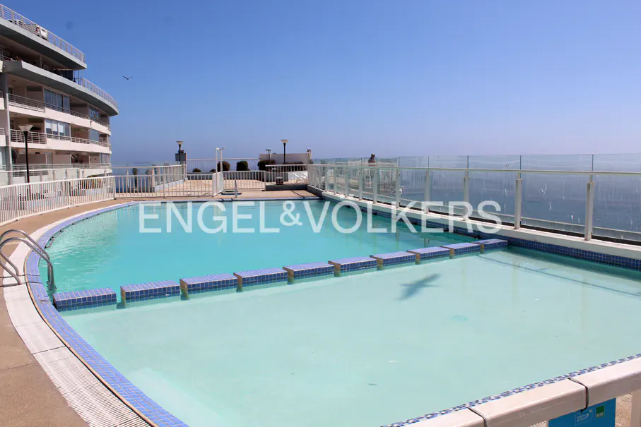Rooftop pool with blue tiles and clear water, overlooking the ocean. A modern building is visible in the background.