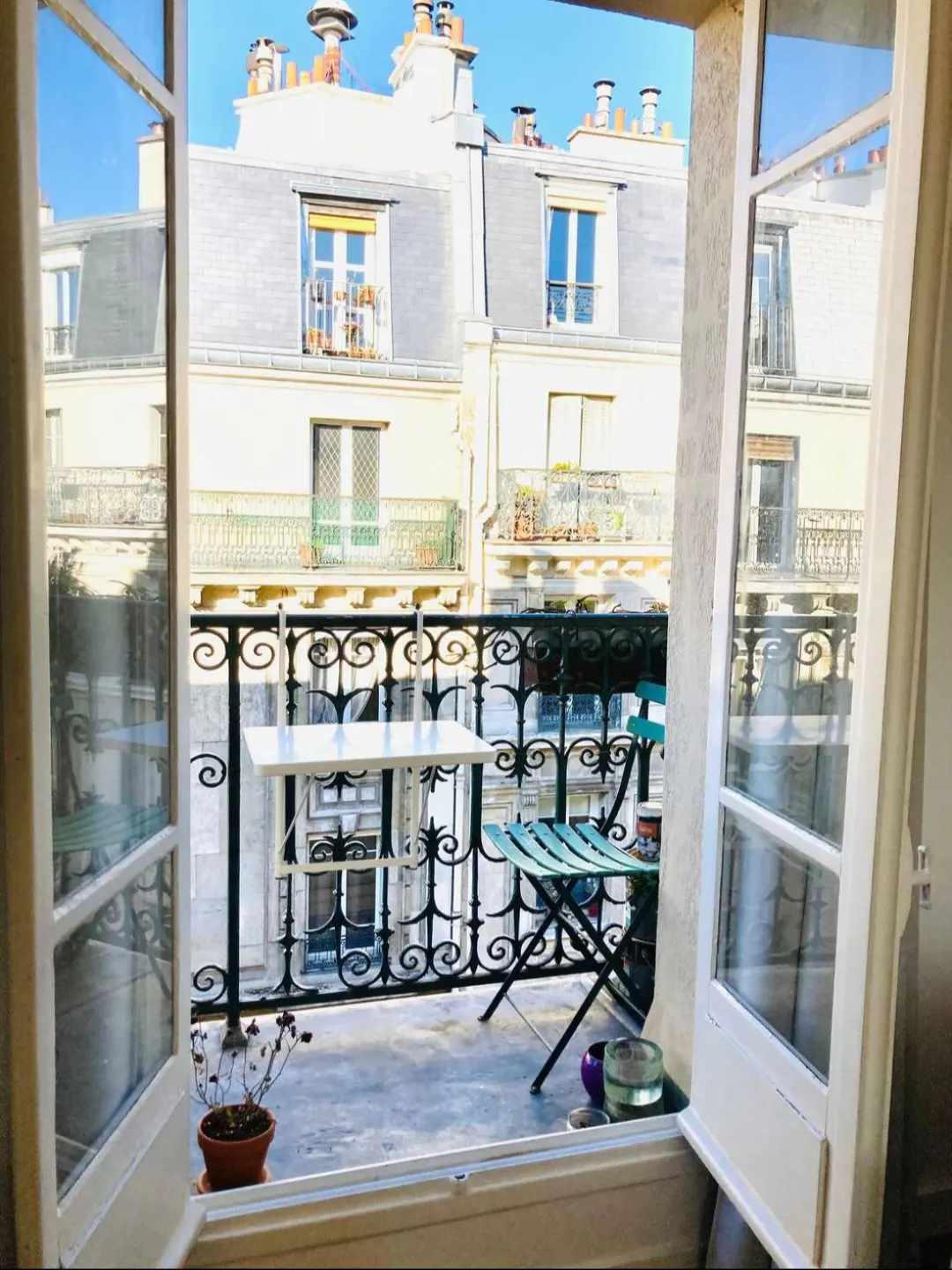 View from open French doors onto a balcony with a table and chair, overlooking Parisian buildings.