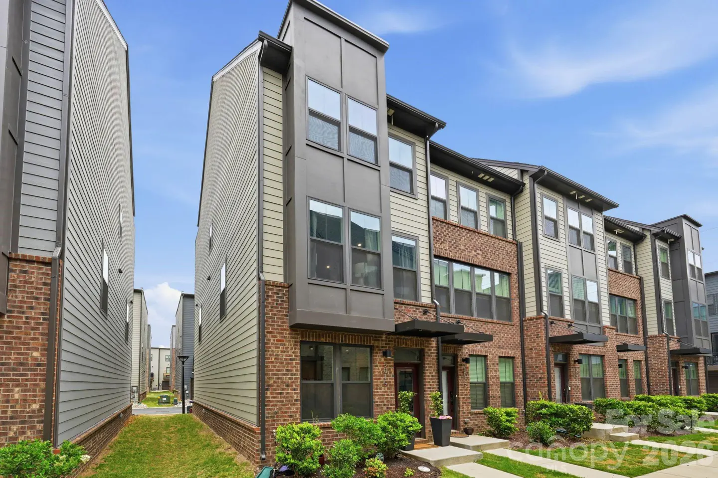 Exterior view of modern townhouses with brick and siding, under a blue sky. Green bushes line the front.