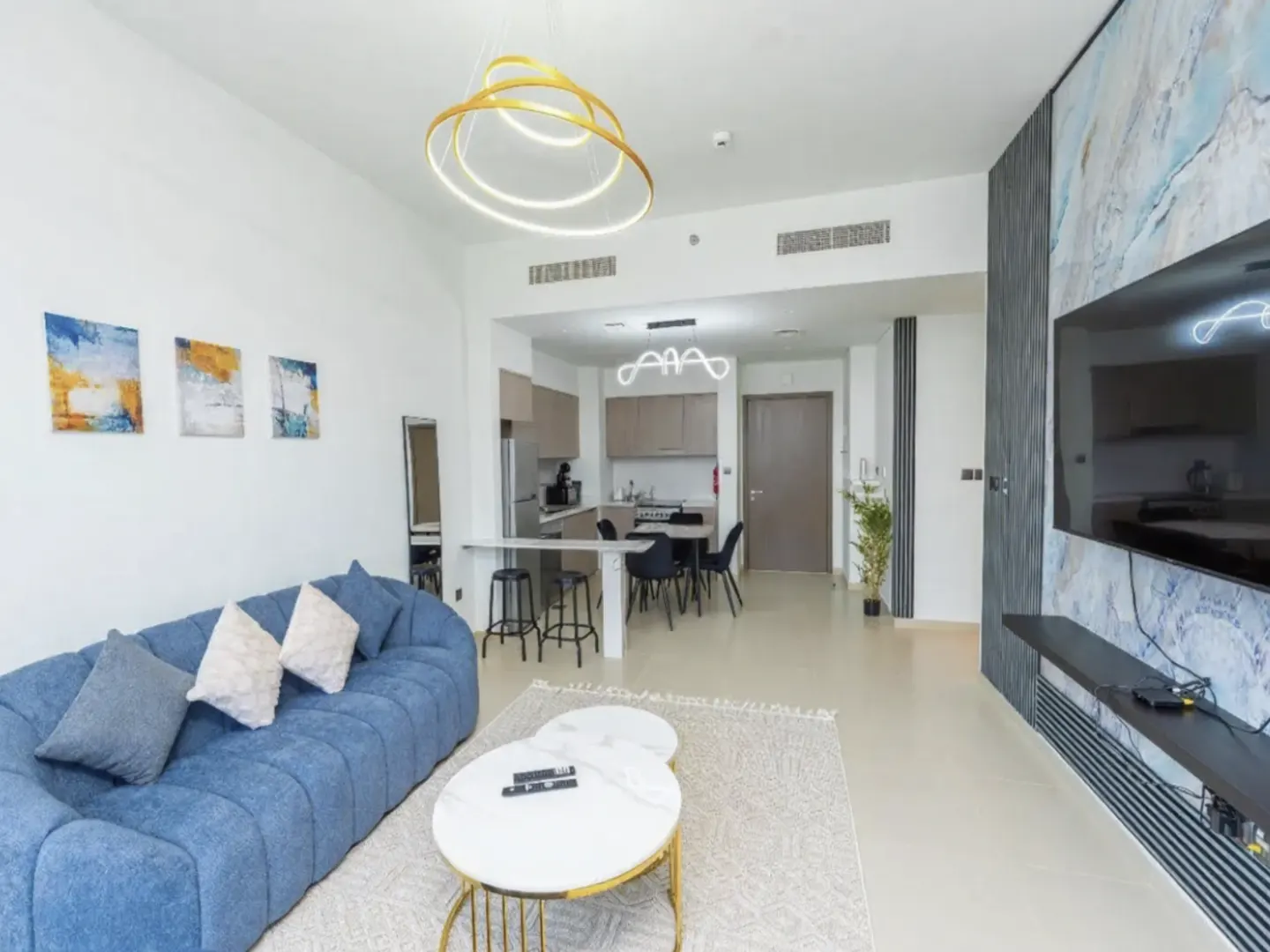 Bright, modern apartment interior with a blue sofa, white walls, and an open kitchen. A gold ring chandelier hangs above the living area.
