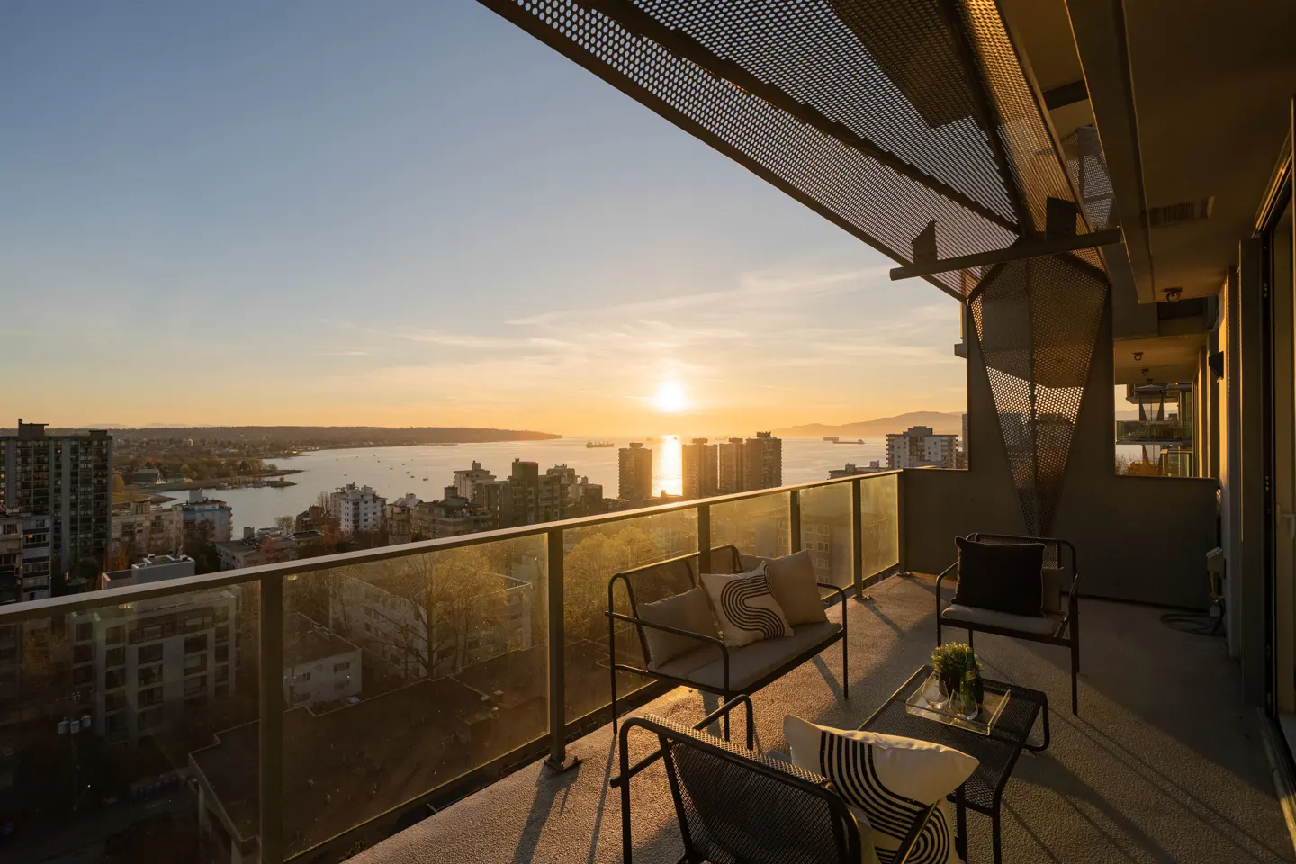 Balcony view at sunset. Black metal furniture with cushions. Glass railing overlooks city, ocean, and setting sun.