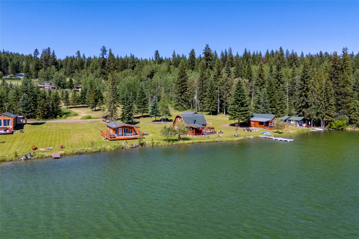 Aerial view of lakefront cabins with green roofs and wood siding, surrounded by lush green trees under a clear blue sky.