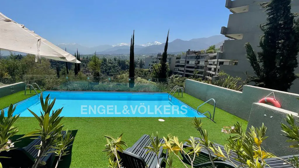 Outdoor pool with blue water, surrounded by green turf, lounge chairs, and a mountain view. Engel & Völkers logo in the pool.