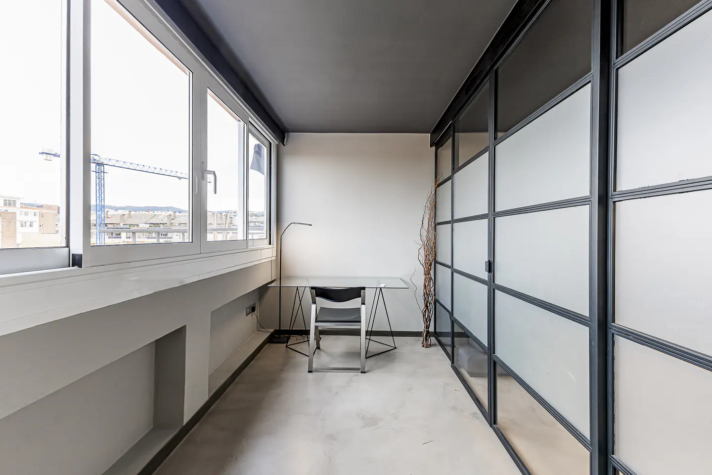 Bright office space with large windows, a desk, and a black-framed frosted glass wall. A city view with a crane is visible.