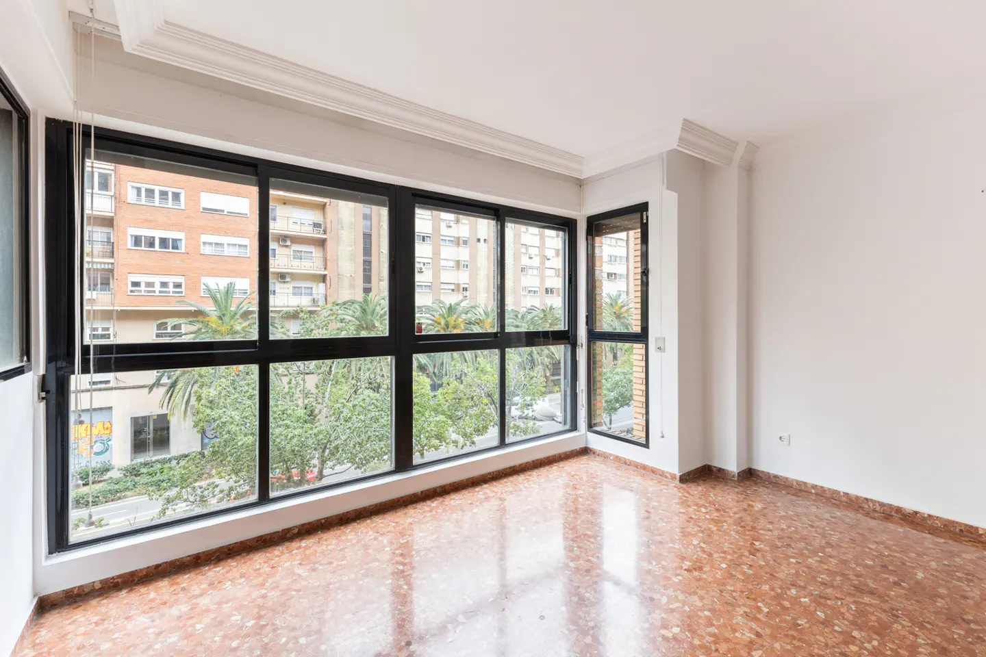 Bright, empty room with large black-framed windows showing city views with trees and buildings. Red terrazzo floor and white walls.