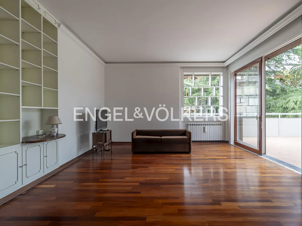 Bright living room with hardwood floors, white walls, and built-in shelving. A brown sofa sits near a window and sliding glass doors.