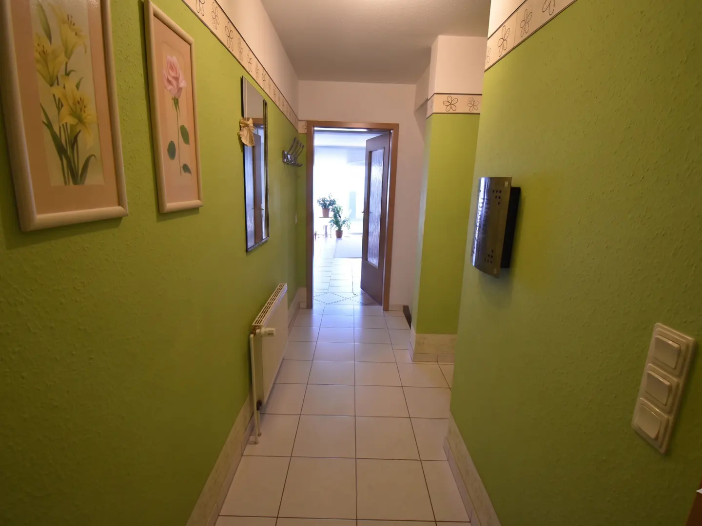 A hallway with bright green walls, white tiled floors, floral wall art, a radiator, and a doorway leading to a bright room.
