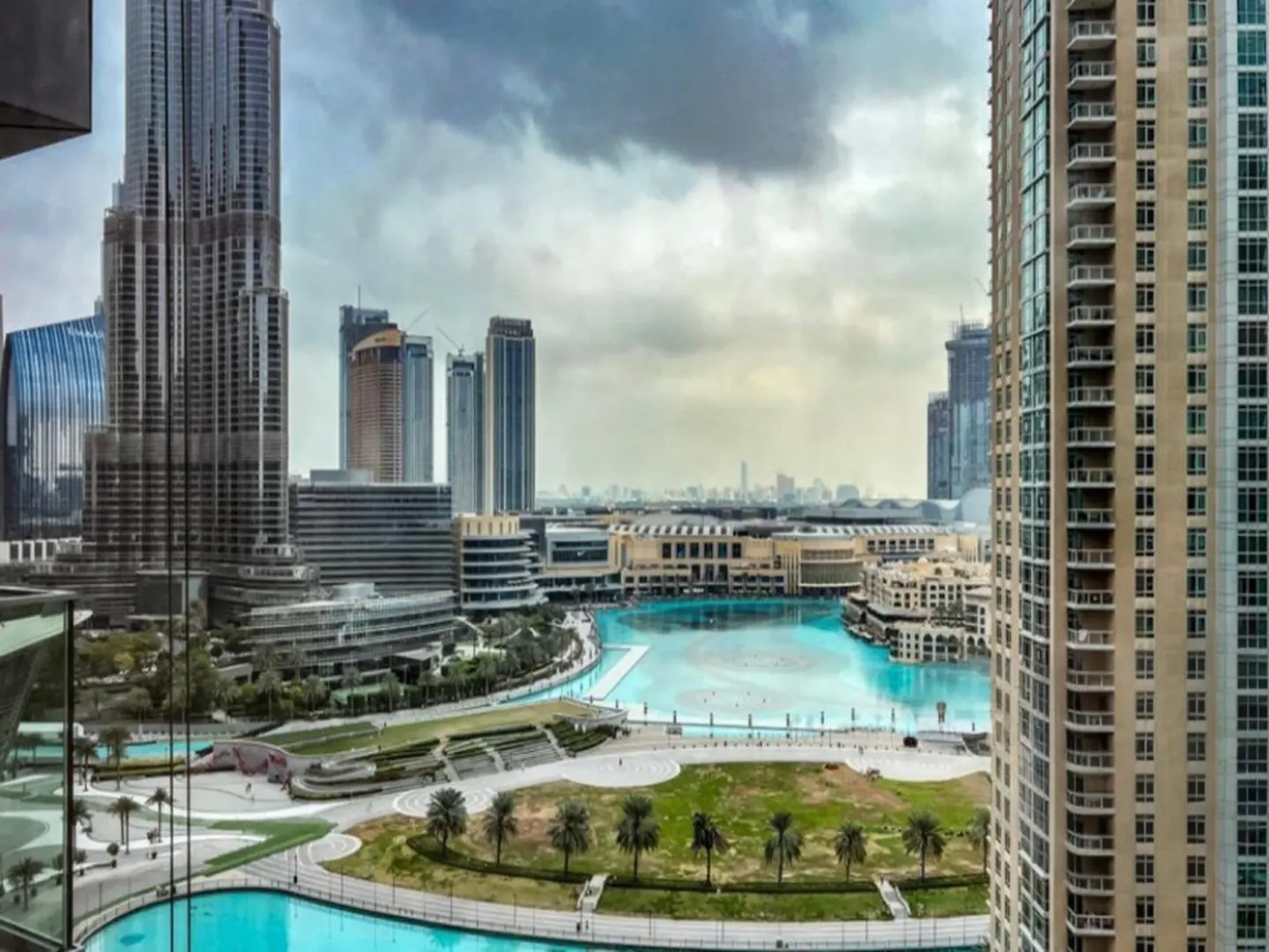 Dubai skyline view from a high-rise, featuring the Burj Khalifa, a turquoise fountain, palm trees, and modern architecture under a cloudy sky.