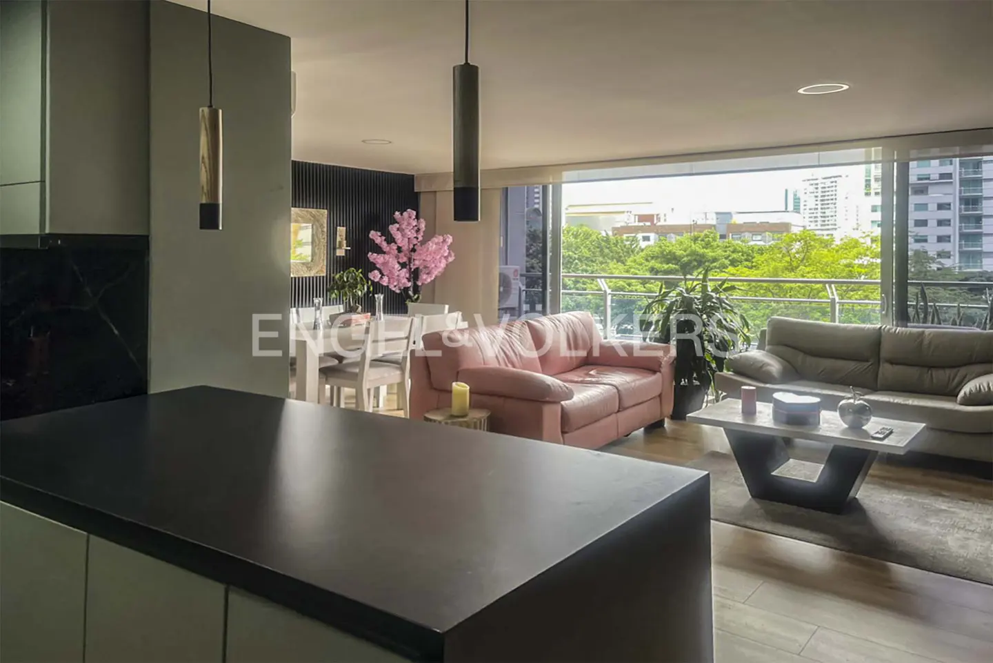 Modern open-plan living area with a dark kitchen island, pink sofa, grey couch, and large windows overlooking city trees.