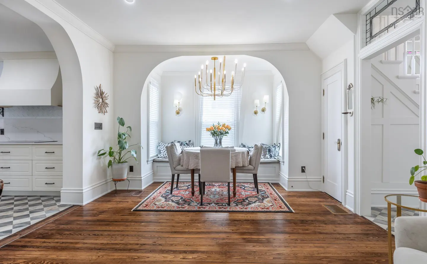 Bright dining room with hardwood floors, white walls, and arched doorway. A round table sits on a patterned rug beneath a gold chandelier. A window seat is in the background.
