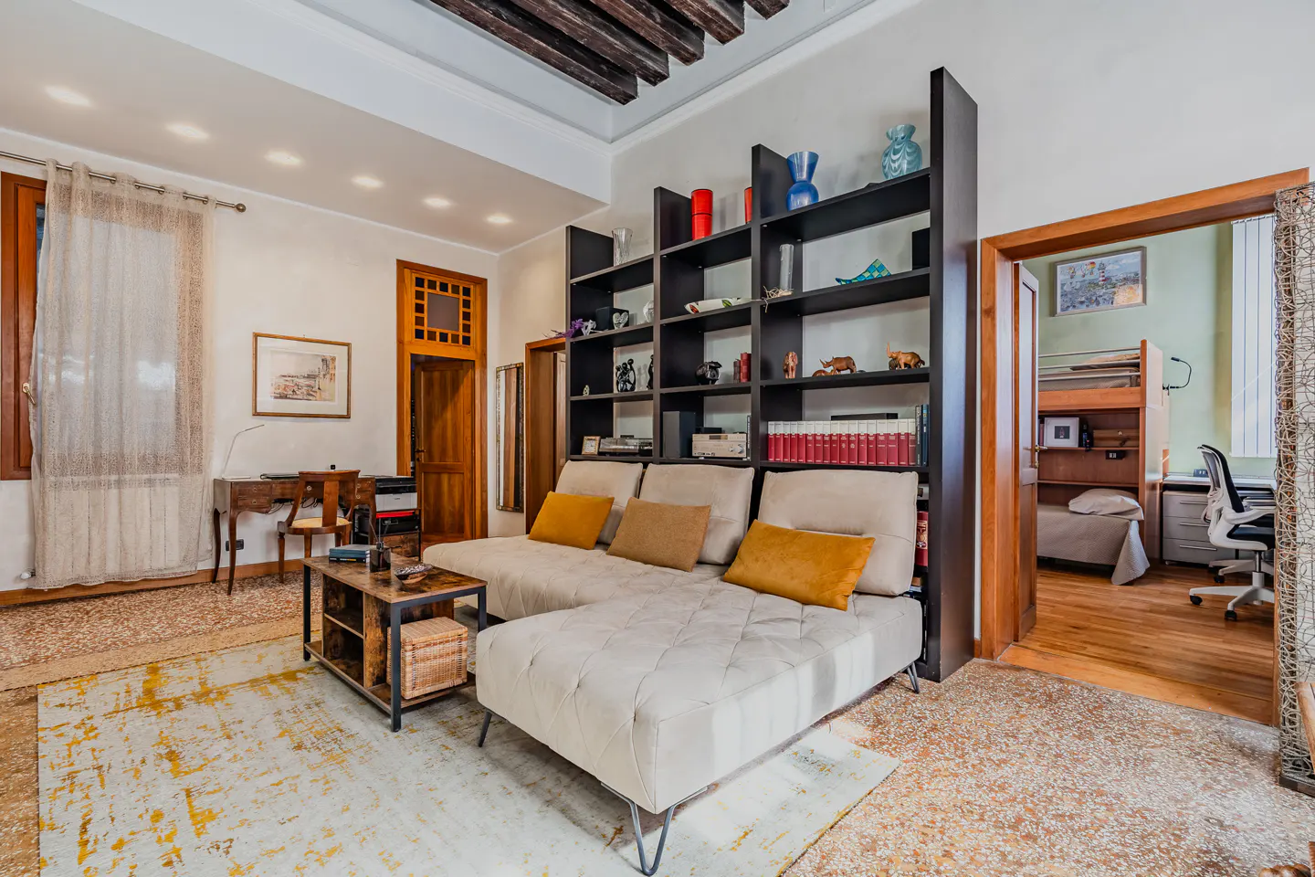 Living room with a beige sectional sofa, dark wood bookshelf, and a view into a bedroom.