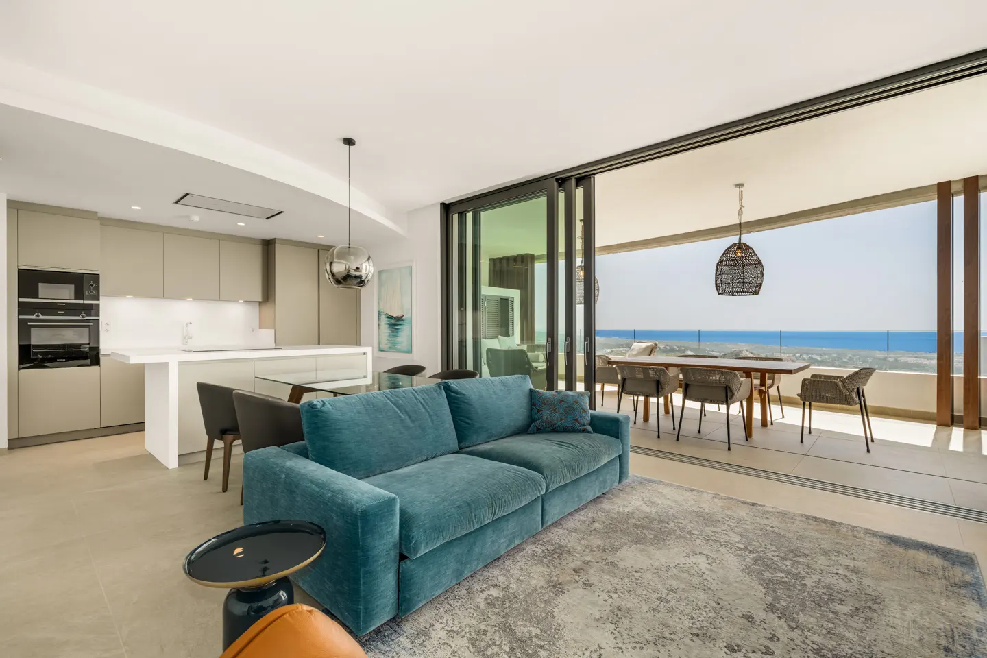 Bright, open-concept living space with a teal sofa, kitchen, and dining area leading to a balcony with ocean views.