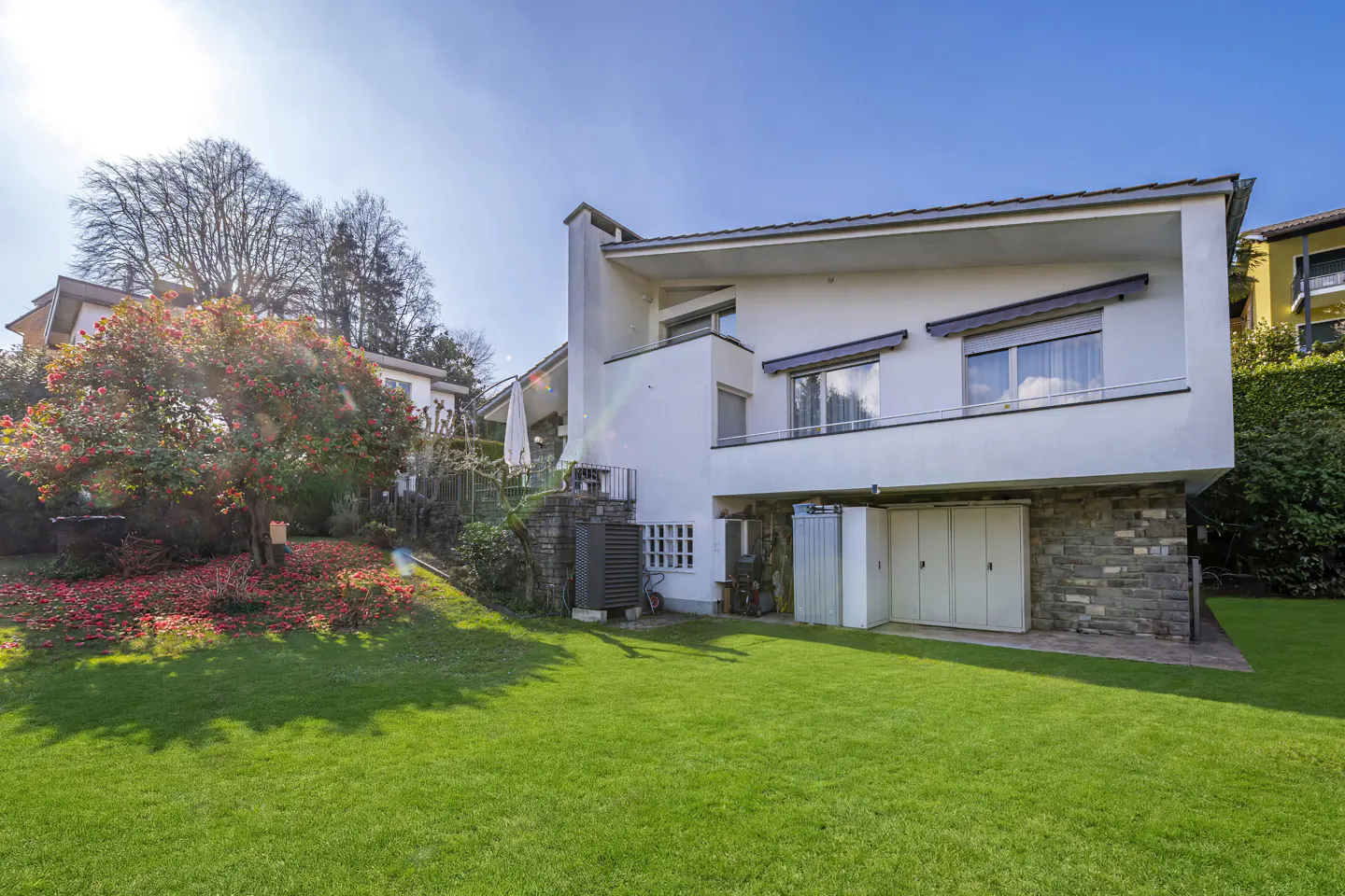 Back view of a modern white house with a green lawn, stone accents, and a blooming red tree.