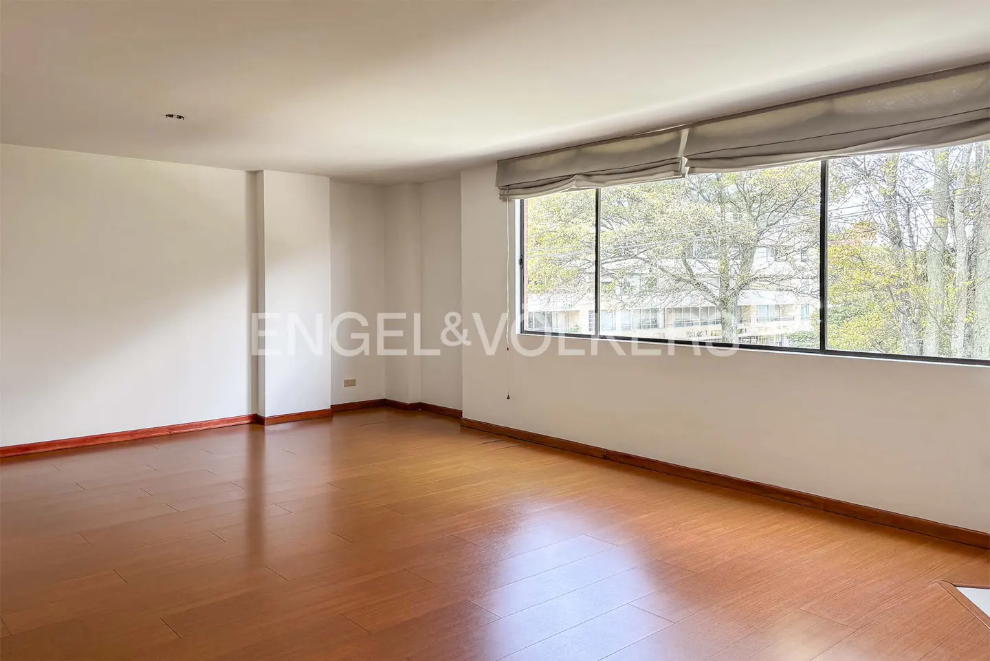 An empty room with hardwood floors, white walls, and a large window with a view of trees. The Engel & Volkers logo is visible.