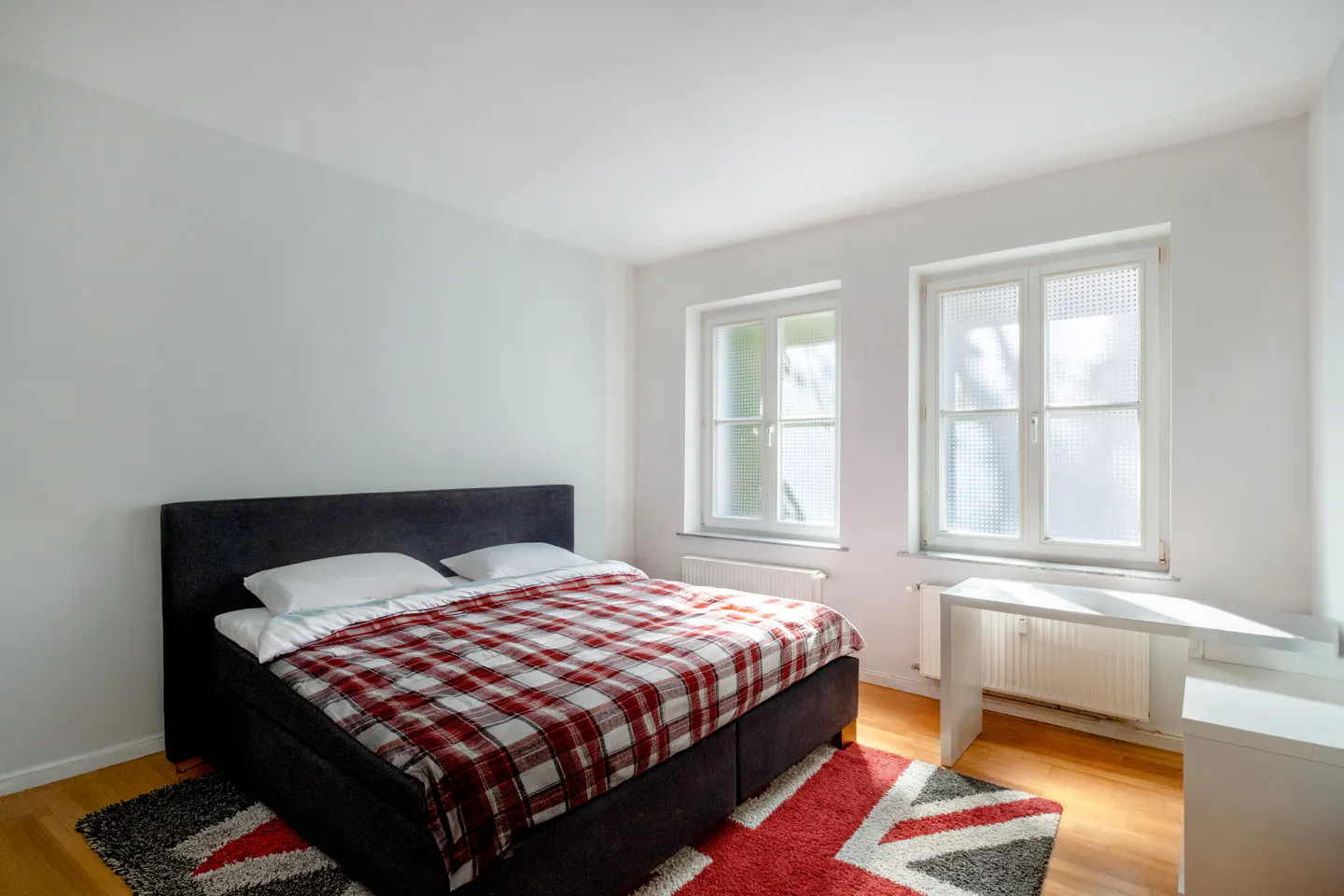 Bright bedroom with a black bed frame, red plaid blanket, and white pillows. Two windows and a white desk are on the right. A red rug is on the floor.