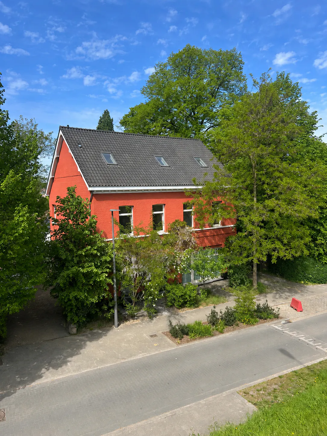 A two-story orange brick house with a dark tiled roof, surrounded by lush green trees under a bright blue sky.