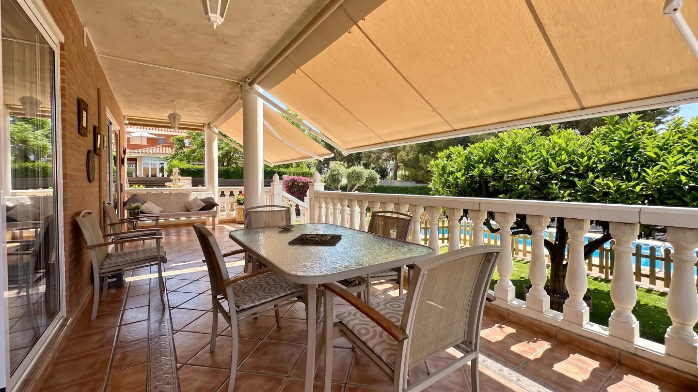 Covered patio with a glass dining table, chairs, and a beige awning overlooking a garden and swimming pool.