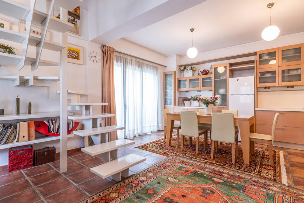Open-plan dining area with a white staircase, wooden table, beige chairs, and colorful patterned rugs on tiled floors.