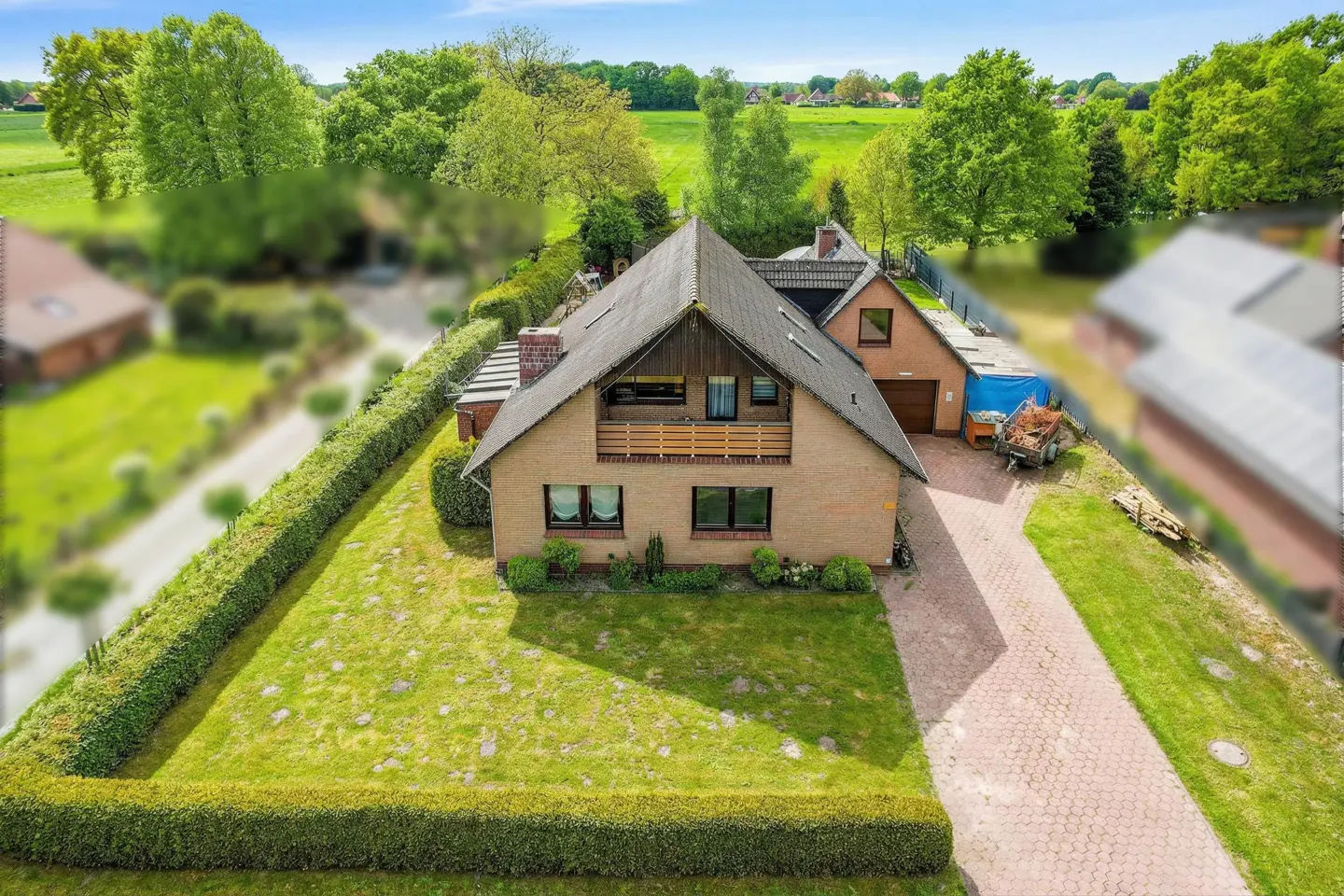 Aerial view of a tan brick house with a dark roof, balcony, and green lawn surrounded by a trimmed hedge. A brick driveway leads to a garage.