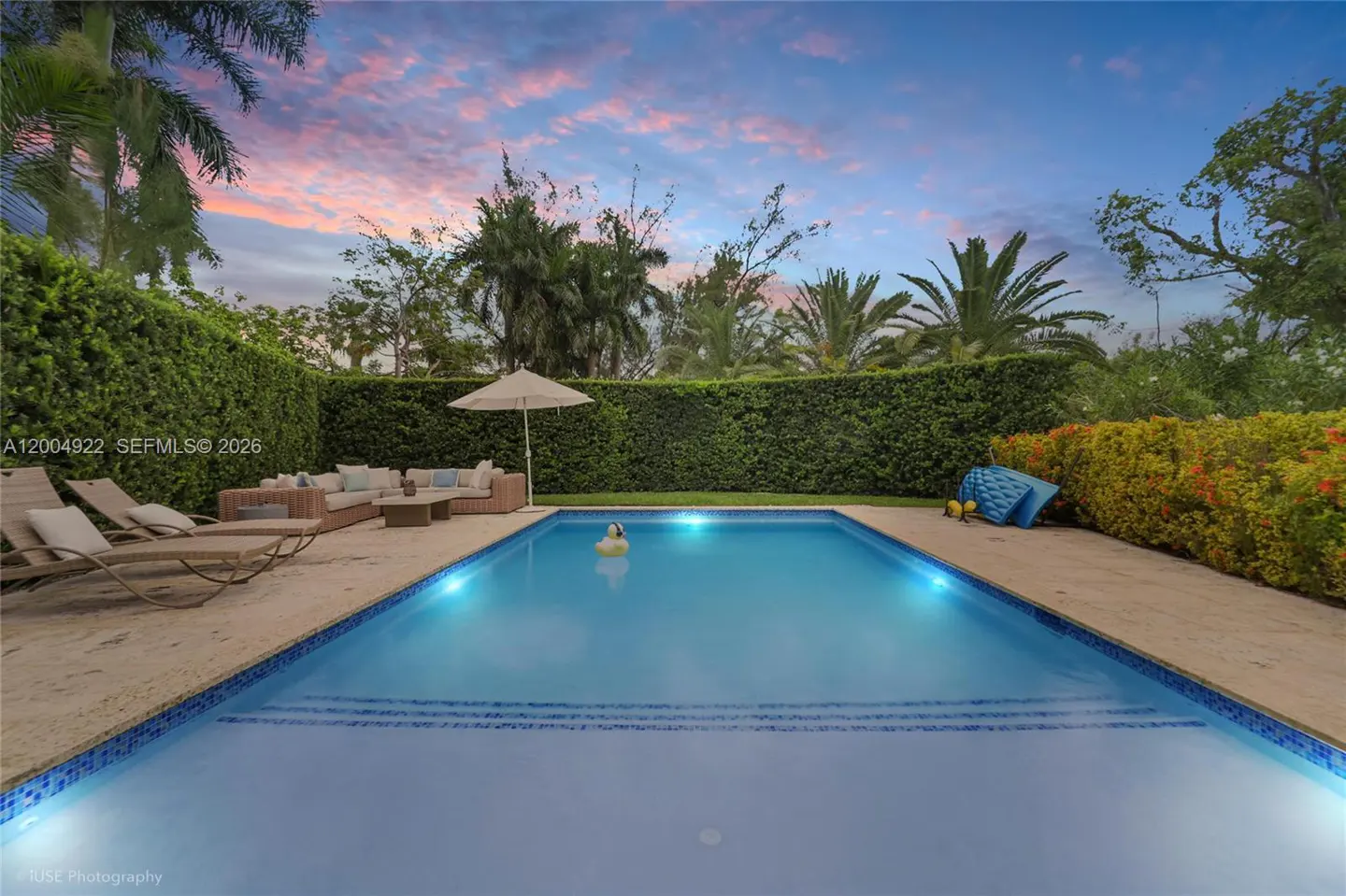Backyard pool with blue water, surrounded by lounge chairs, sofa, and green hedges under a pink and blue sky.