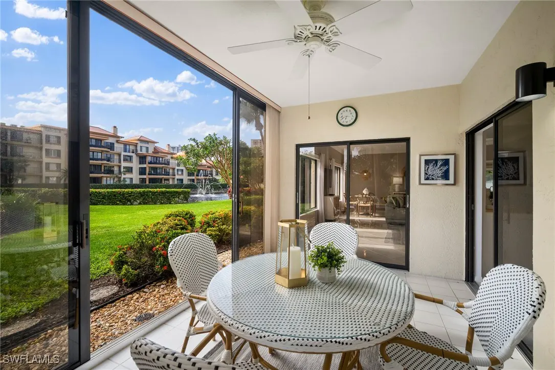 Screened-in porch with white wicker table and chairs. View of green lawn, fountain, and multi-story building through sliding glass doors.