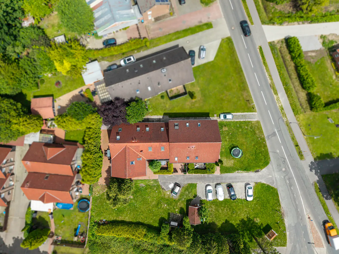 Aerial view of a red-roofed house with cars parked in the driveway, surrounded by green lawns and trees. A road runs alongside the property.