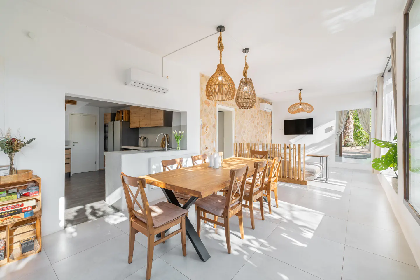 Bright, open-concept dining area with a wooden table, chairs, and woven pendant lights. Kitchen and outdoor view in the background.