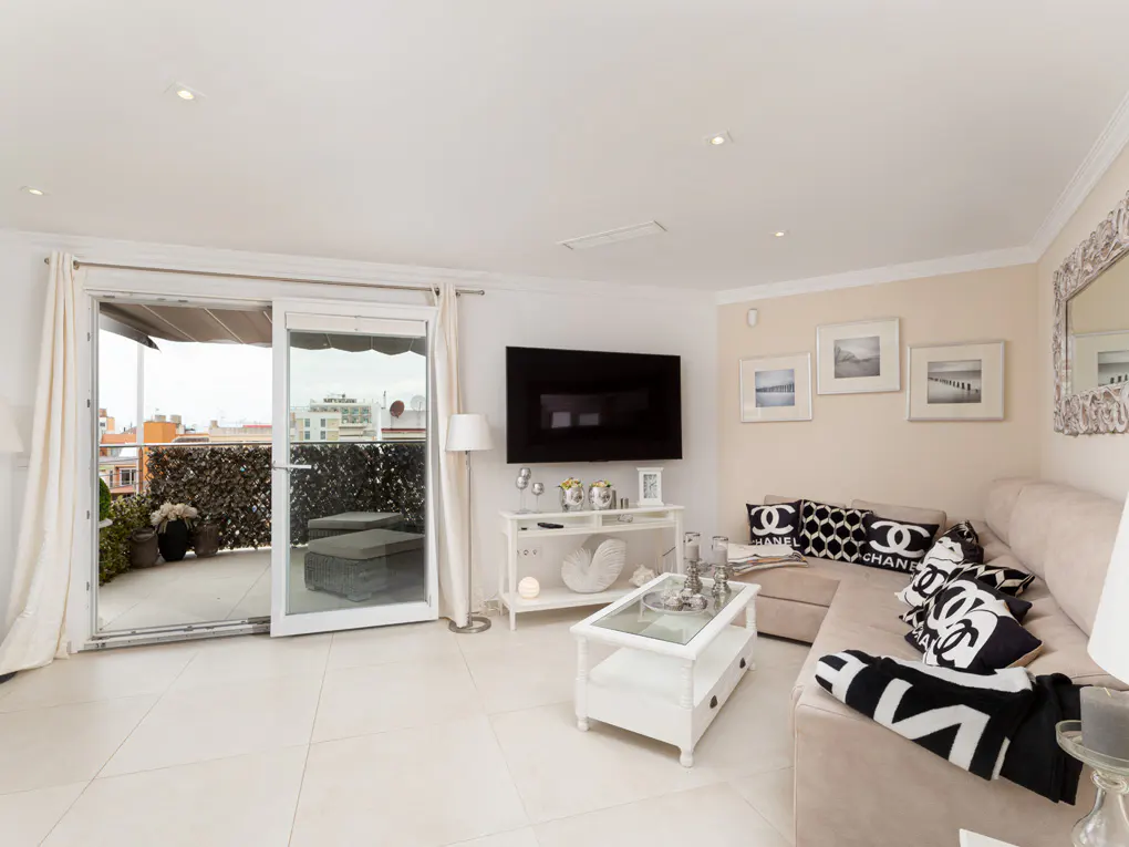 Living room with beige sofa, black and white pillows, and a white coffee table. Sliding glass doors open to a balcony with outdoor seating.