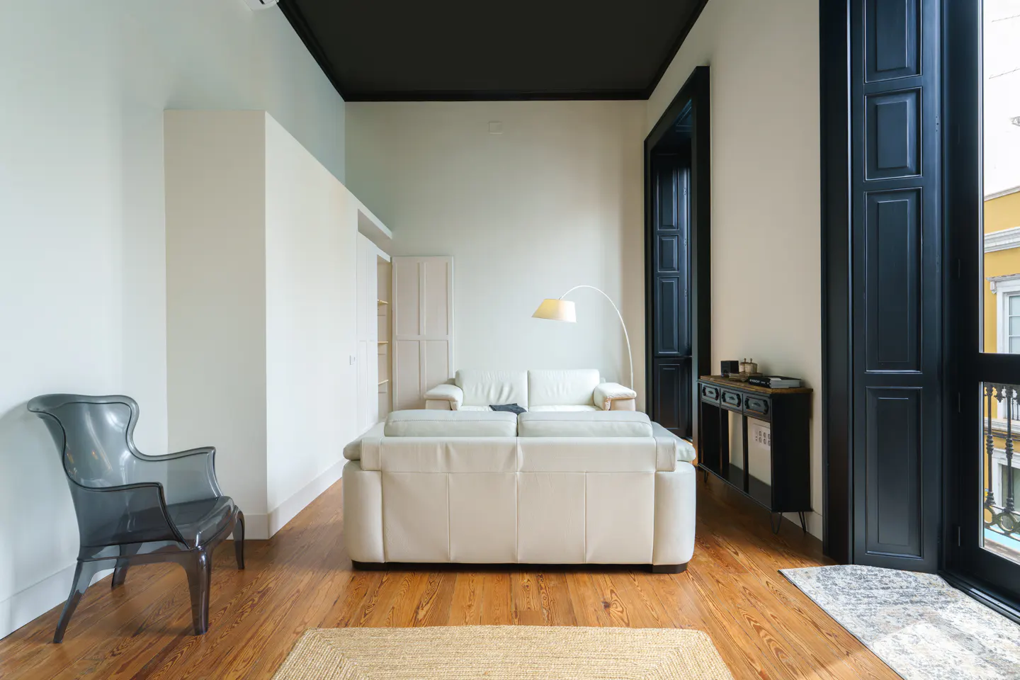 Bright living room with white walls, wood floors, and a white sofa. A black chair sits near a window with black shutters.