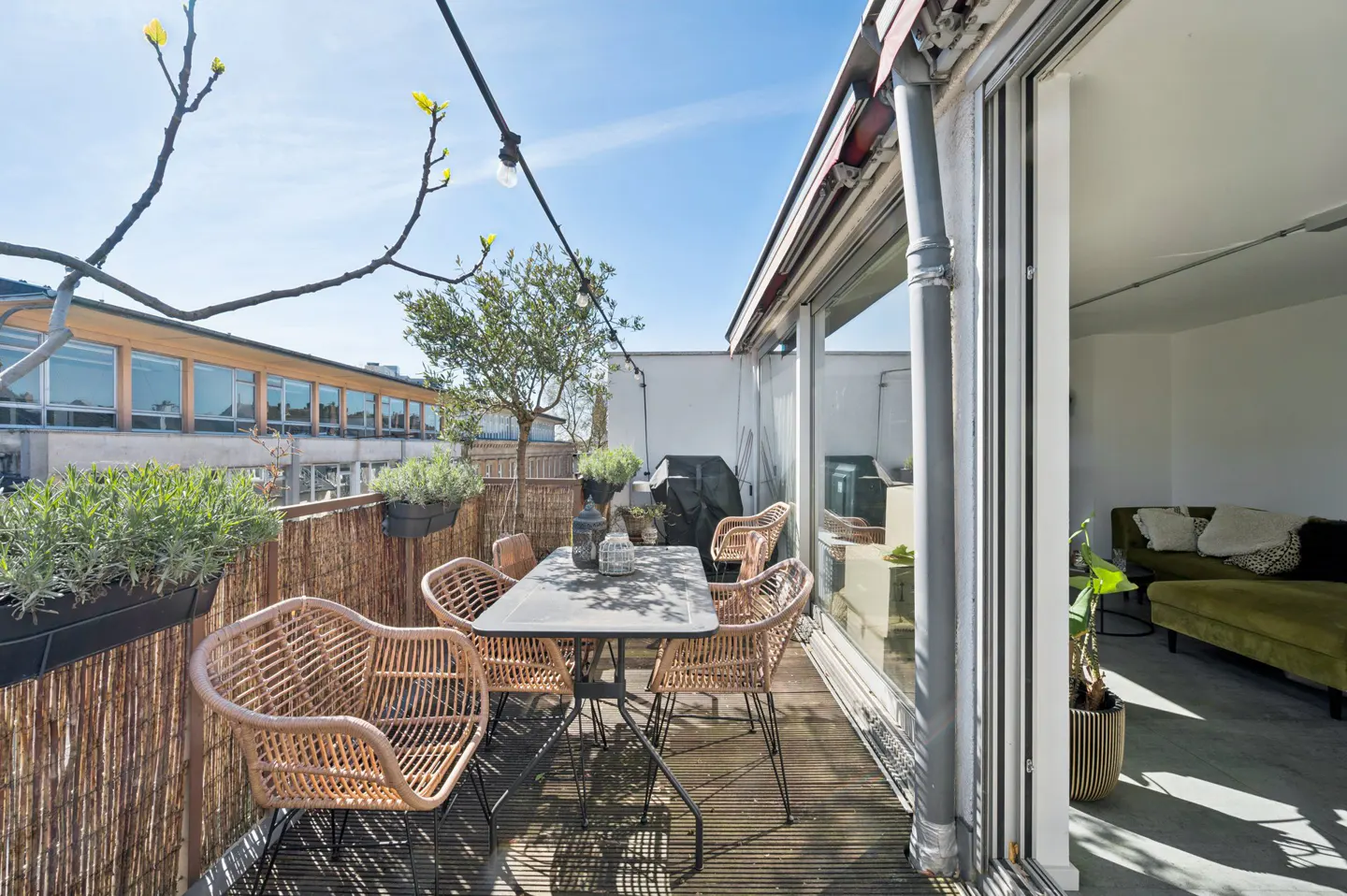 Outdoor patio with a table, wicker chairs, and a bamboo fence with plants. Sliding glass doors lead to a living room with a green sofa.