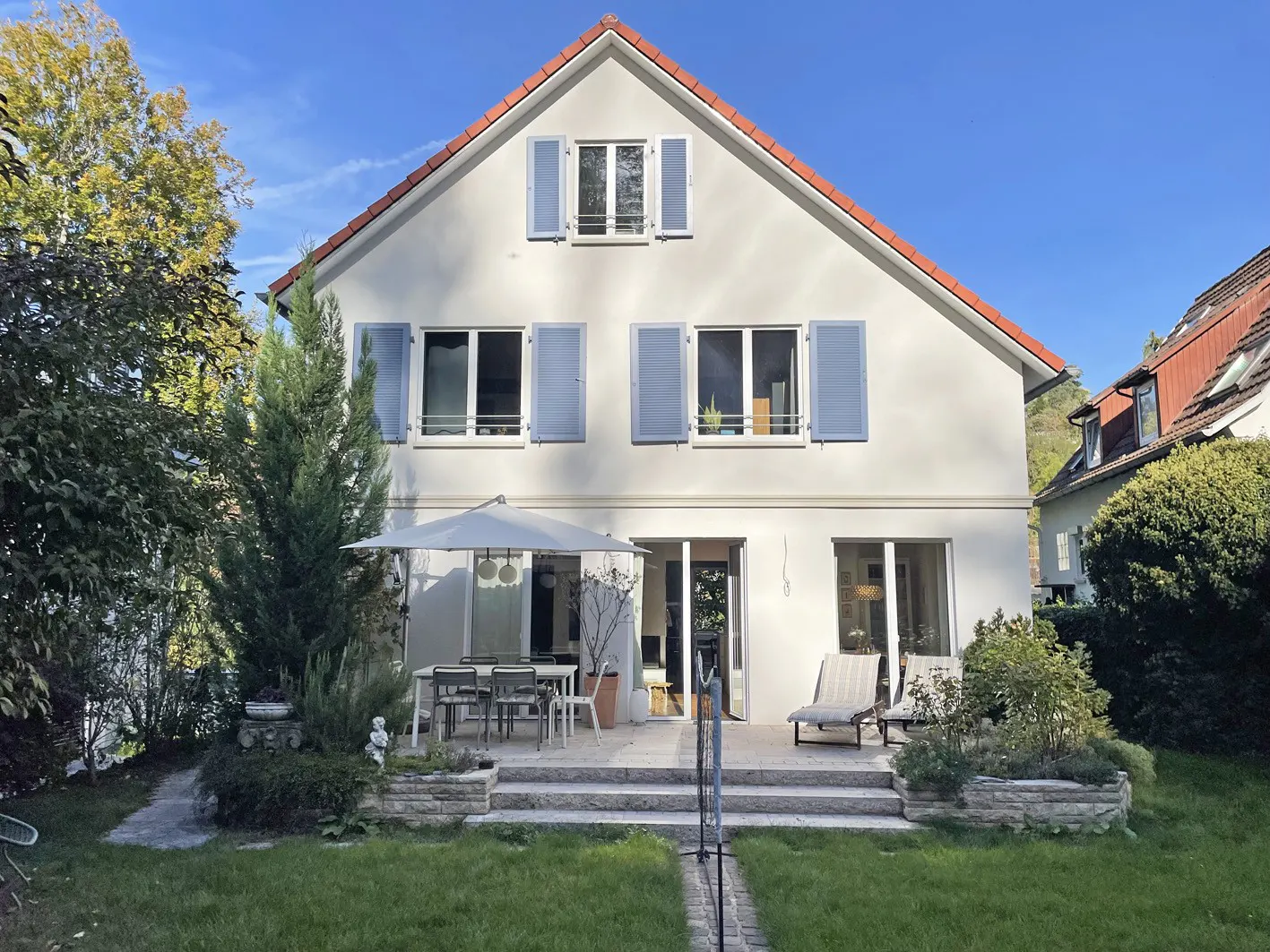 Two-story house with white walls, red roof, and blue shutters. Patio with table, chairs, umbrella, and lounge chairs. Green lawn and trees.
